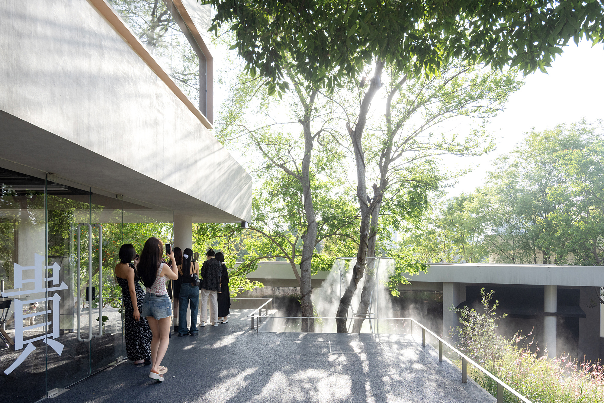 Visitors gather on a paved terrace under tall trees beside a glazed ground floor and a concrete overhang, with mist rising beyond