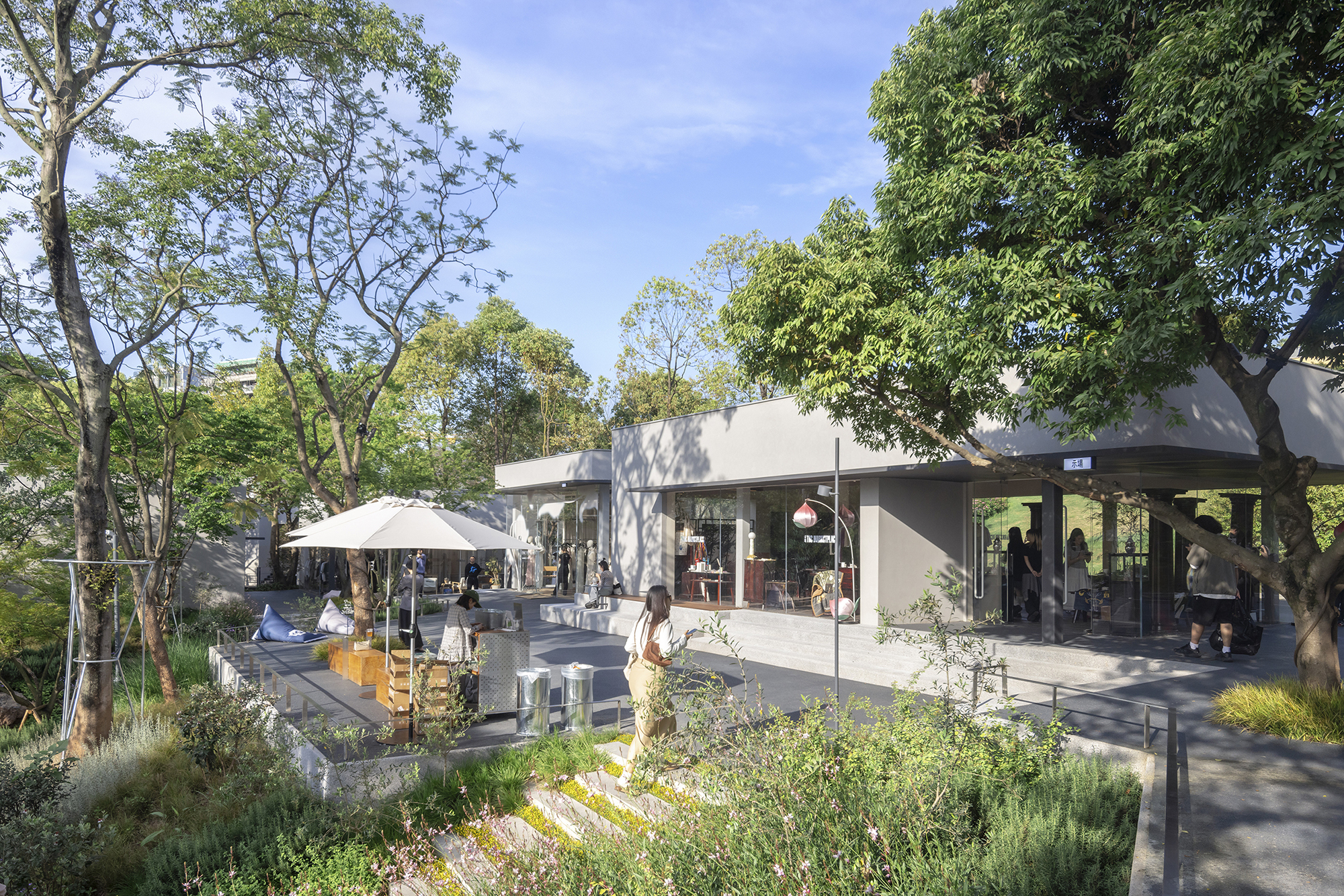 Low glazed pavilion with a flat concrete roof seen across a planted slope, with people and a white umbrella under afternoon sun