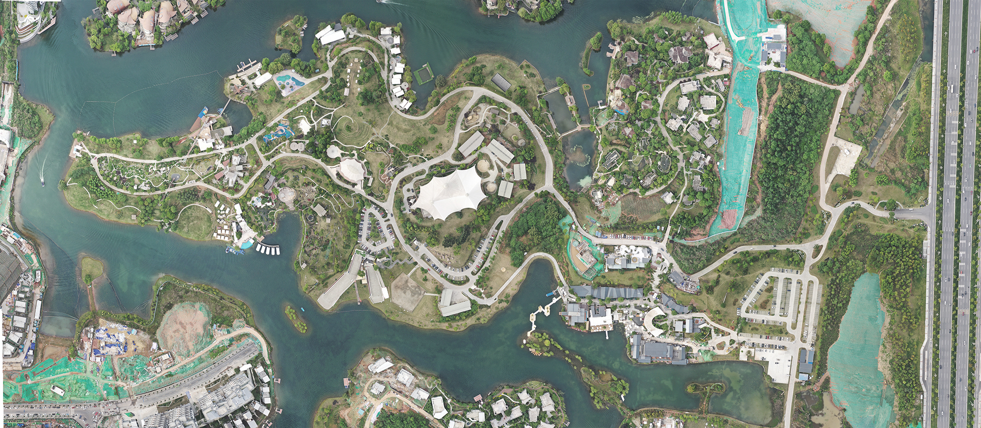 Top-down aerial view of a lakeside site with winding paths, a white tent-like roof at the center and a highway on the right