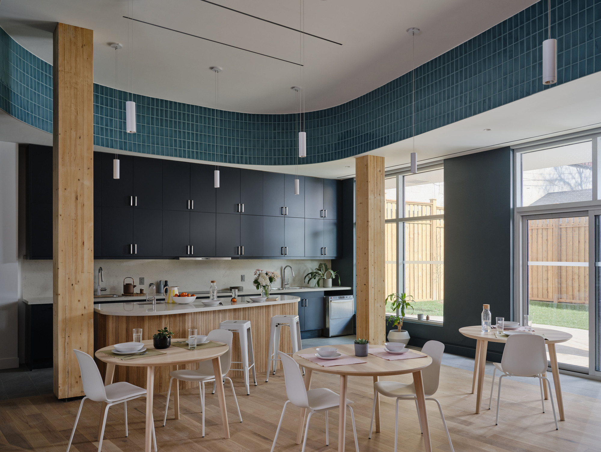Open kitchen and dining area with dark cabinetry, timber columns and a curved teal tile band, lit by daylight from the garden windows