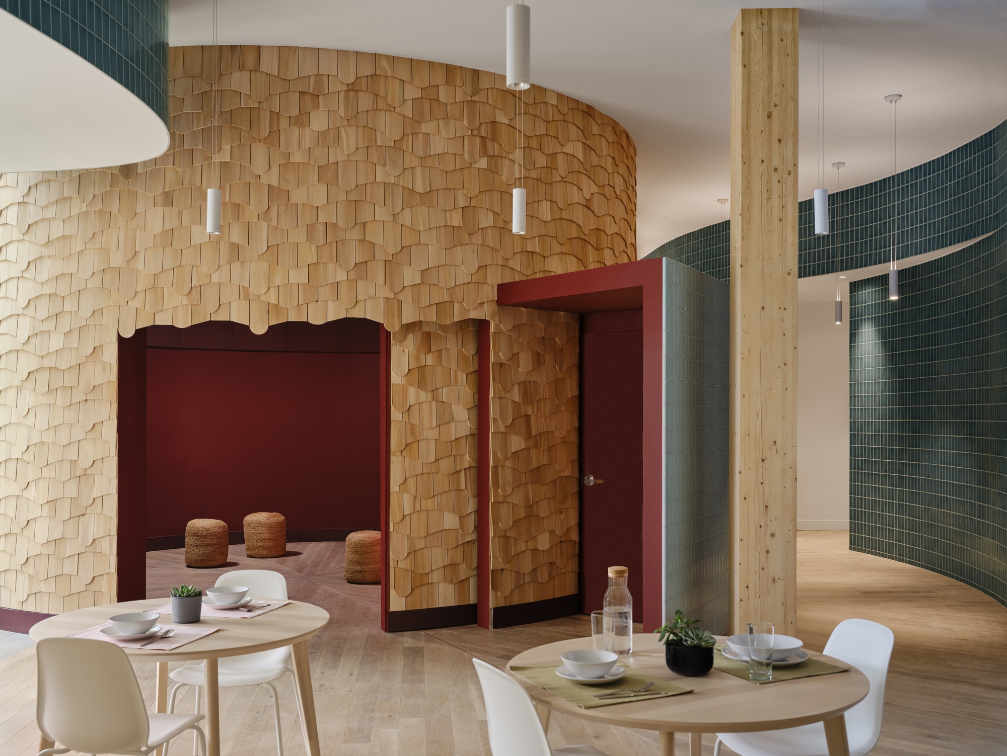 Curved wall clad in layered wood shingles with a red-framed opening, beside a timber column and teal tile wall in soft daylight