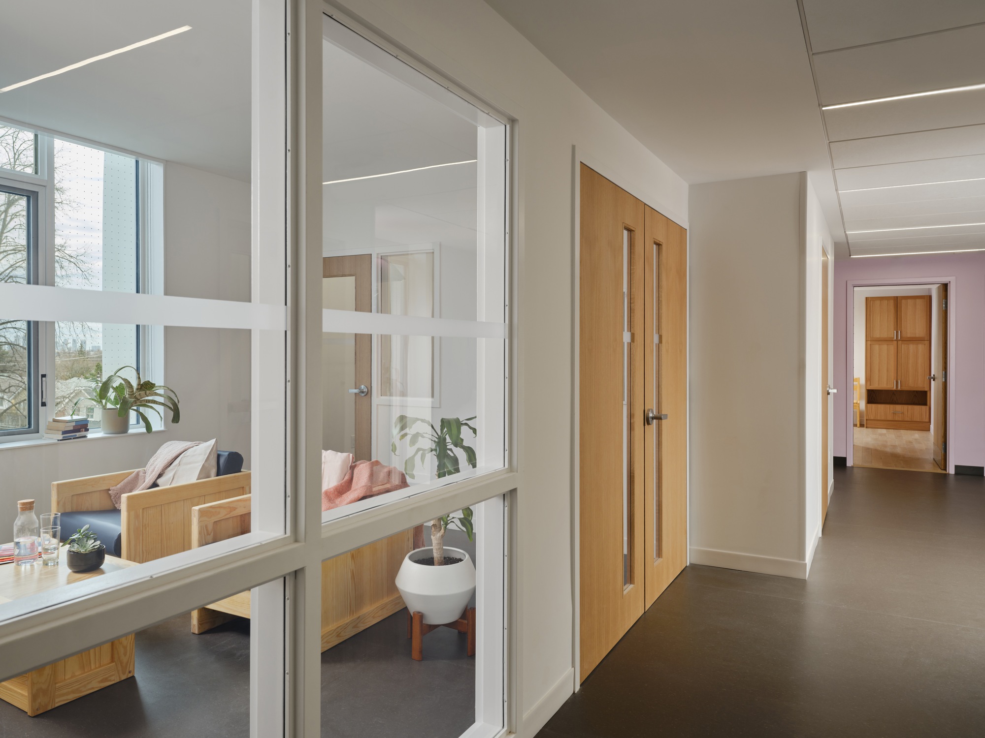Corridor with timber double doors and a glazed partition looking into a sitting room with wooden armchairs and a bare winter tree outside