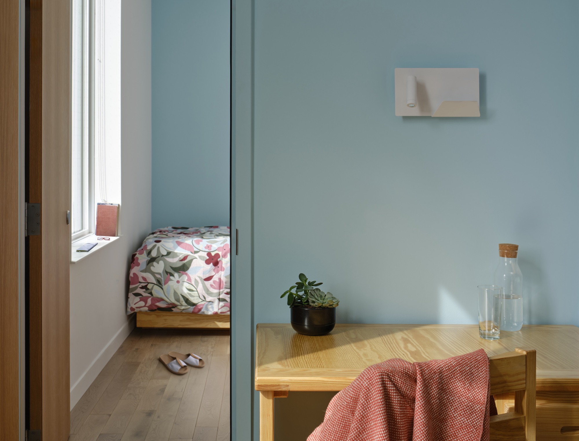 Bedroom corner in pale blue with a wooden desk, glass carafe and succulent, and a floral-covered bed seen through an open door in low sun