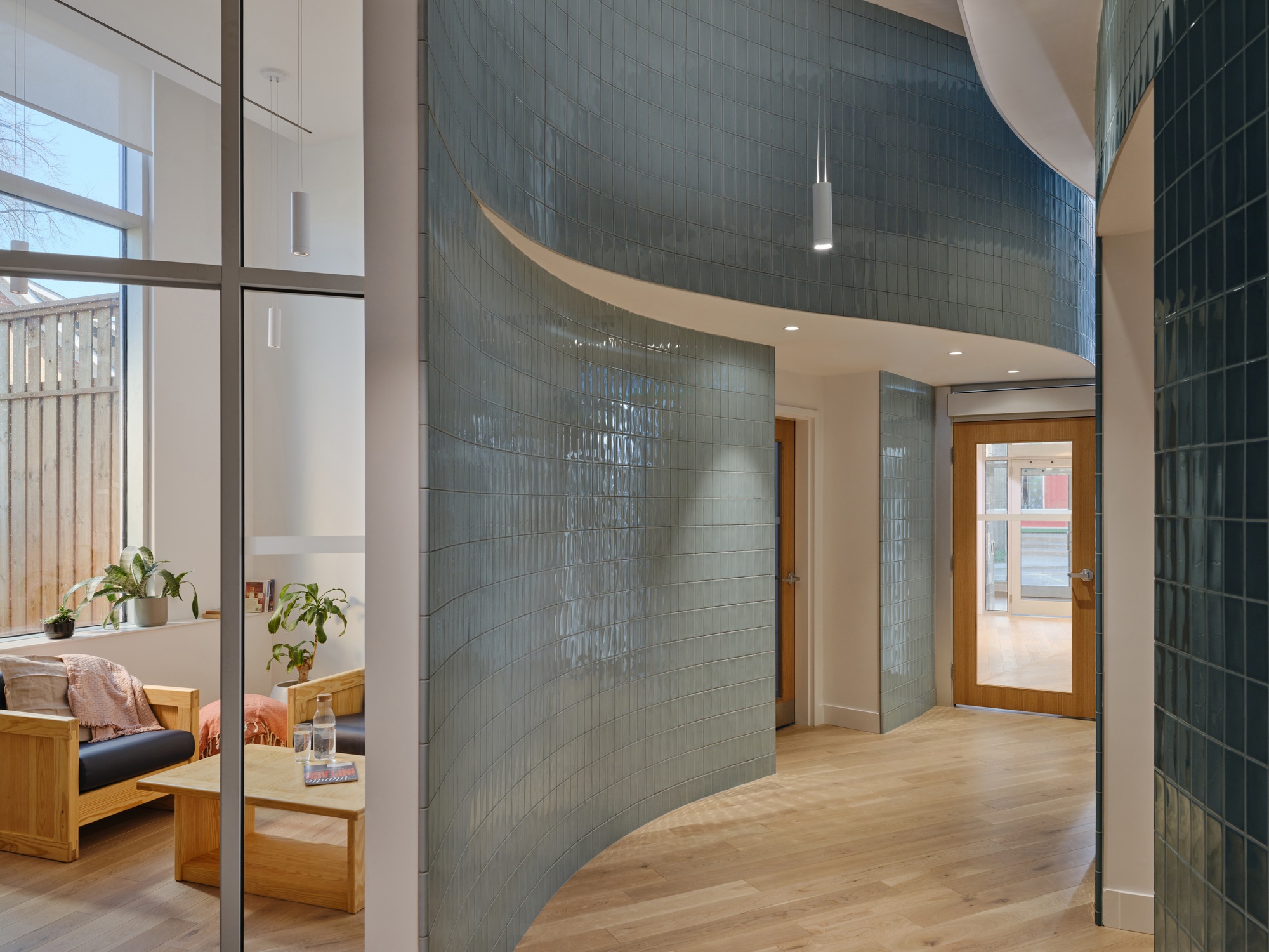 Curving glossy teal tile walls along a timber-floored passage, with a glazed partition opening onto a lounge and a fenced yard