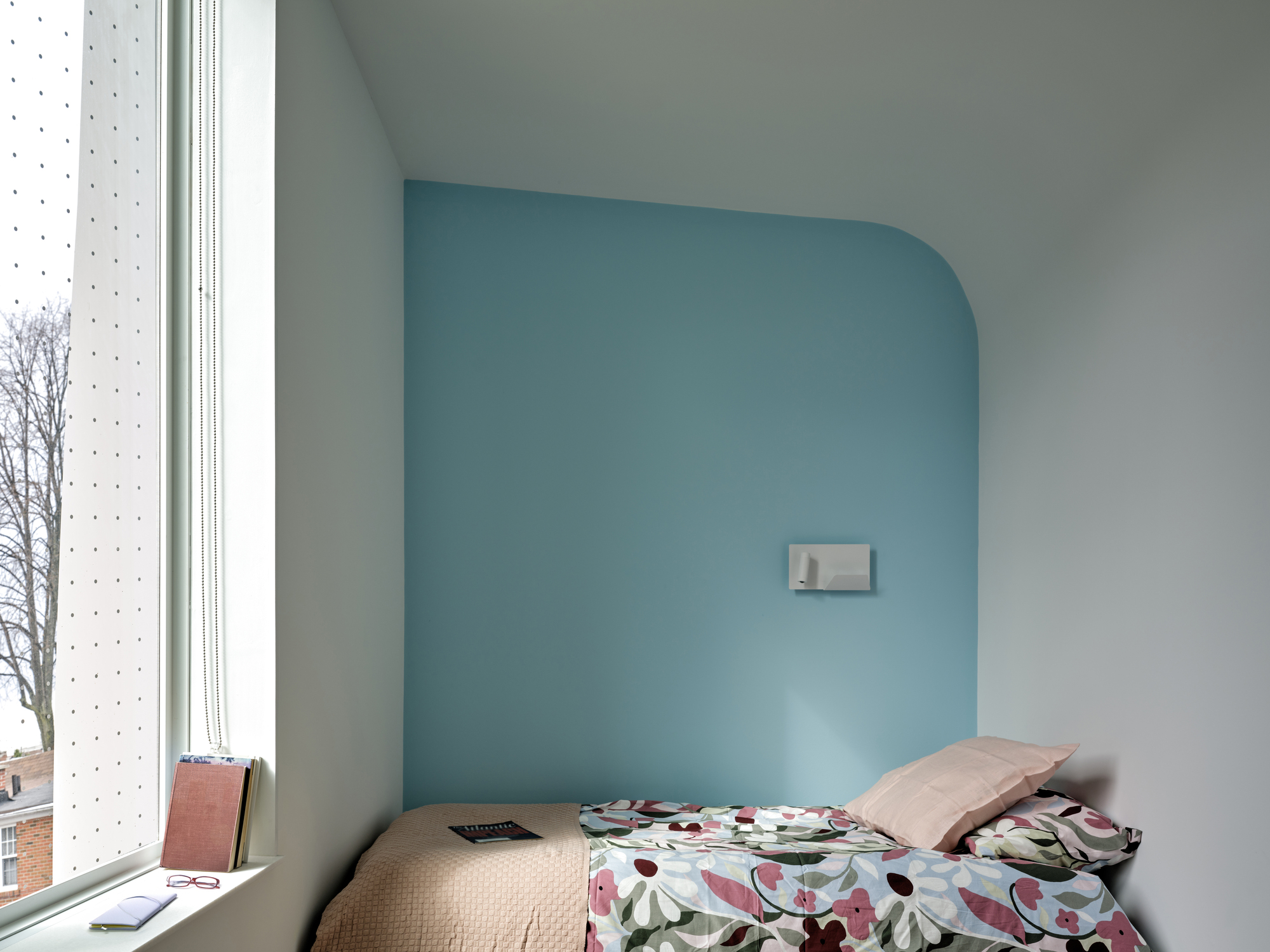 Small bedroom nook with a rounded pale blue wall, floral bedding and a tall perforated window facing a bare tree