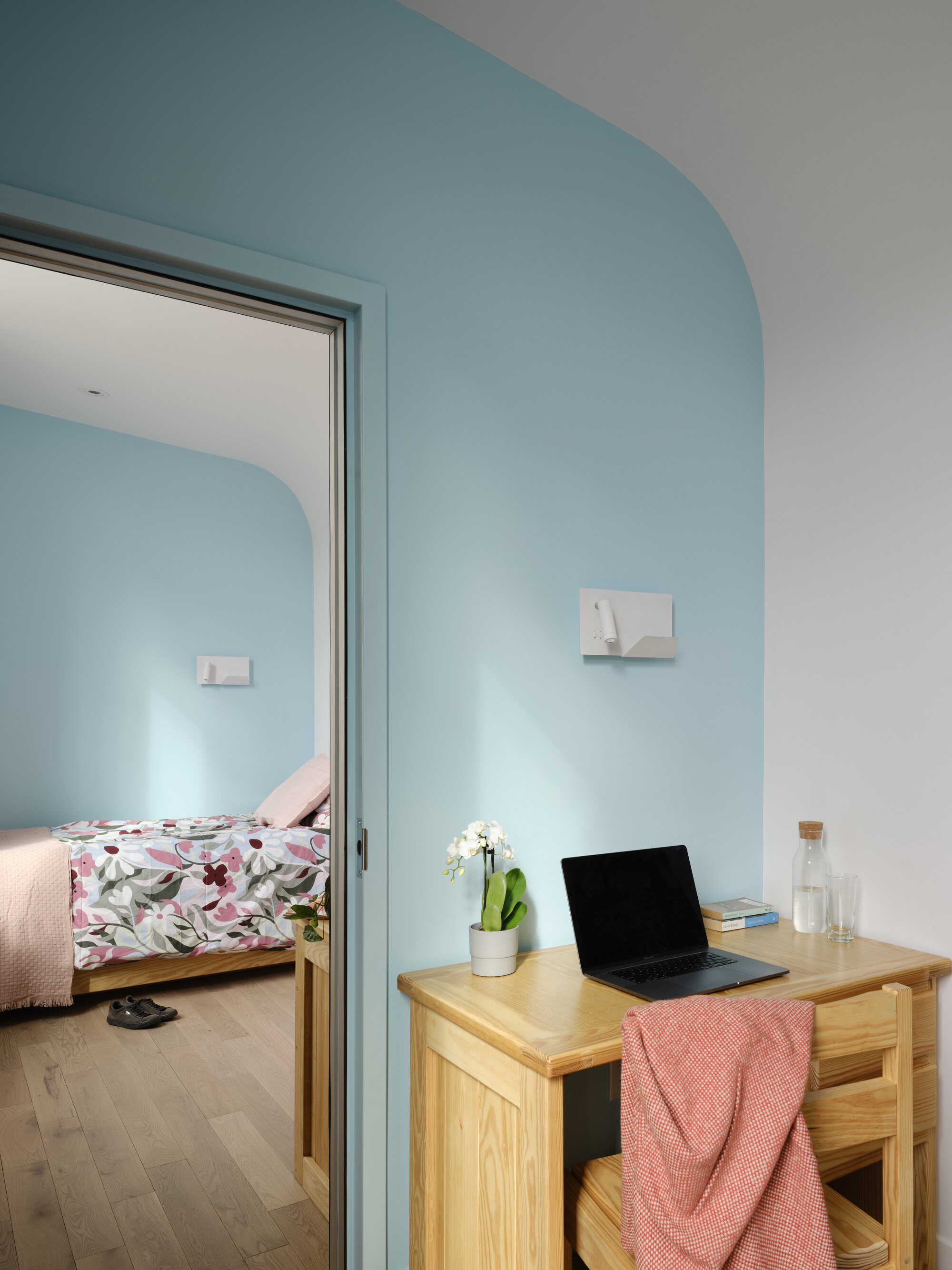 Pale blue curved wall with a timber desk and laptop beside an open door onto a bedroom with floral bedding