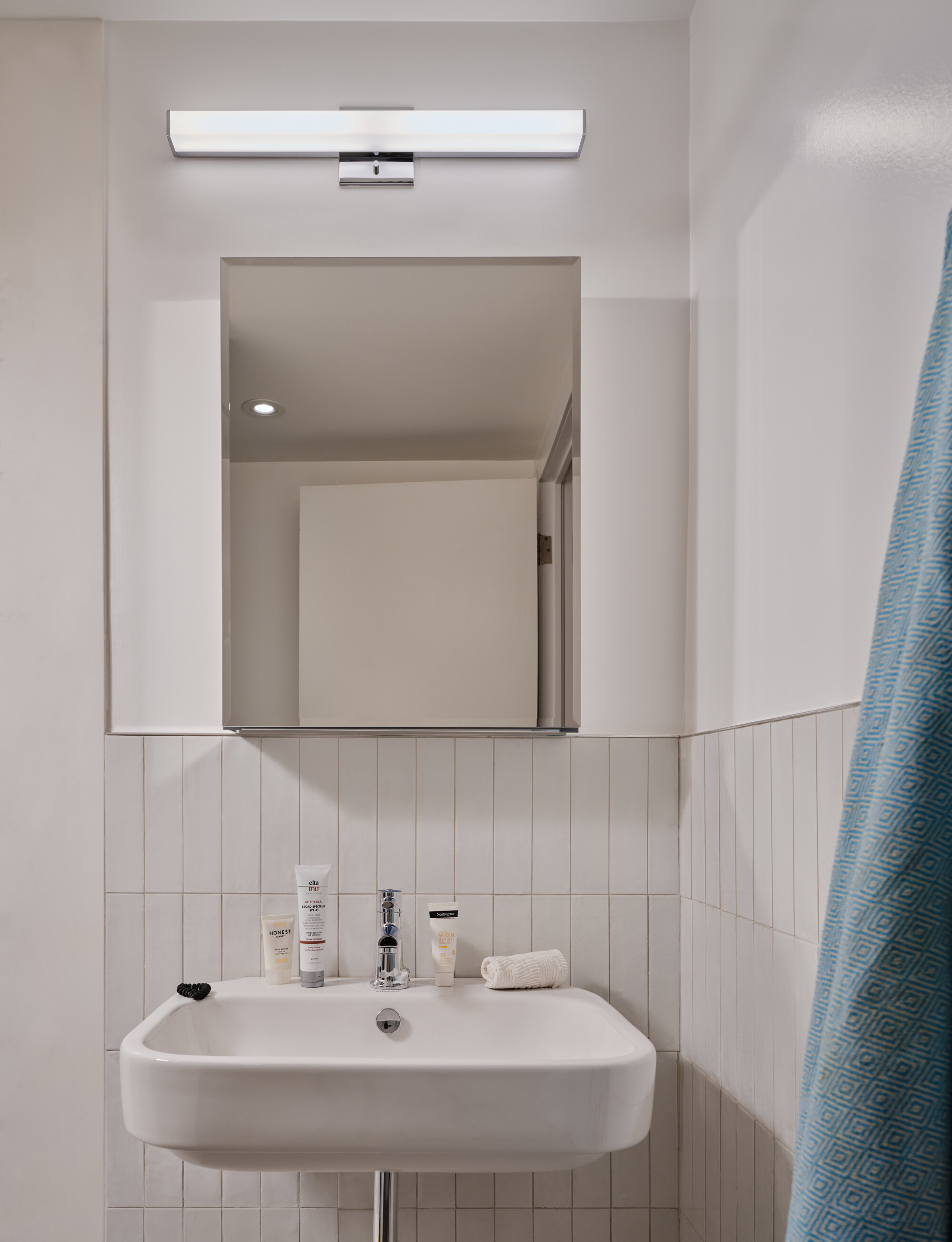 Wall-mounted white basin below a rectangular mirror and strip light, with vertical white tiles and a blue towel at the edge