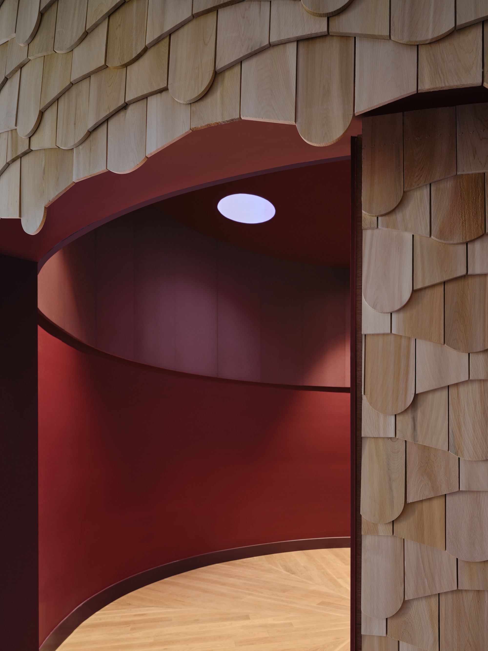 Wood shingle edge framing a doorway into a curved red room with a round skylight and herringbone floor