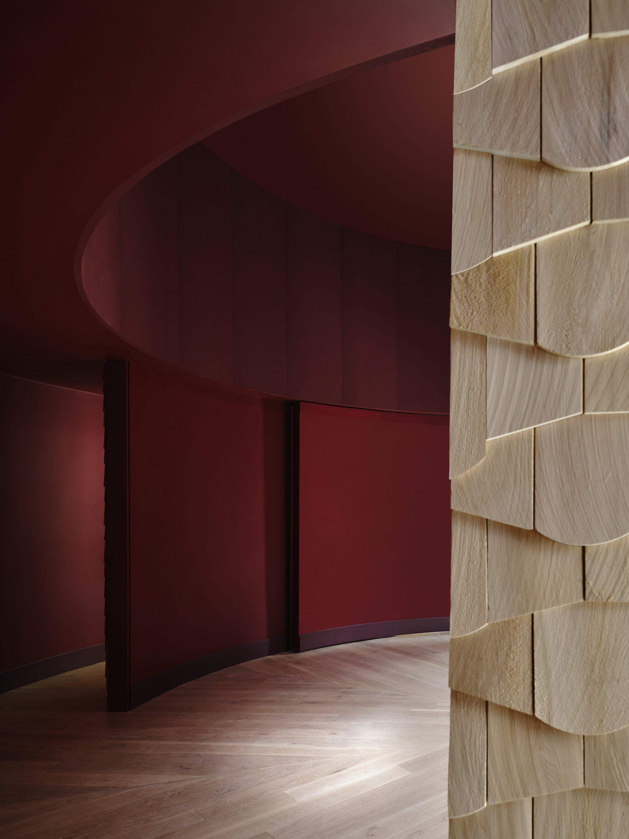 Close-up of a shingle-clad column in the foreground with a curved red wall and herringbone floor beyond in low light