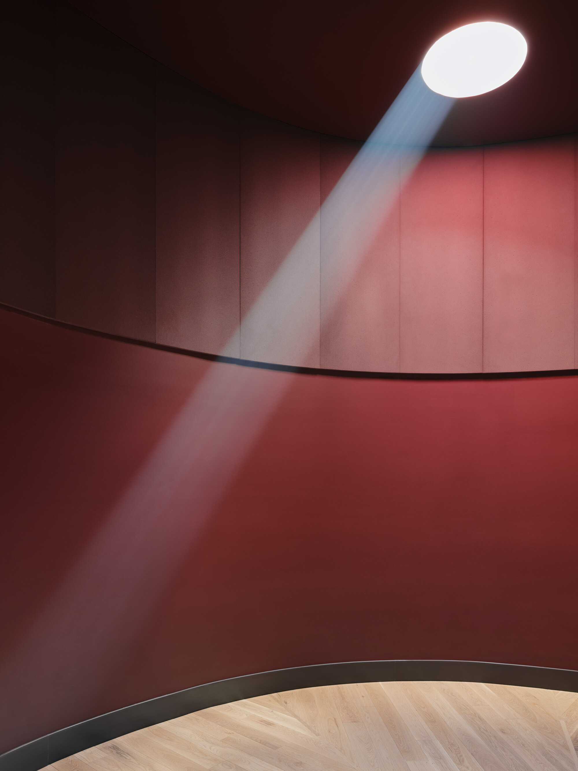 Beam of daylight falling from a round skylight across curved red panelled walls above a pale herringbone floor