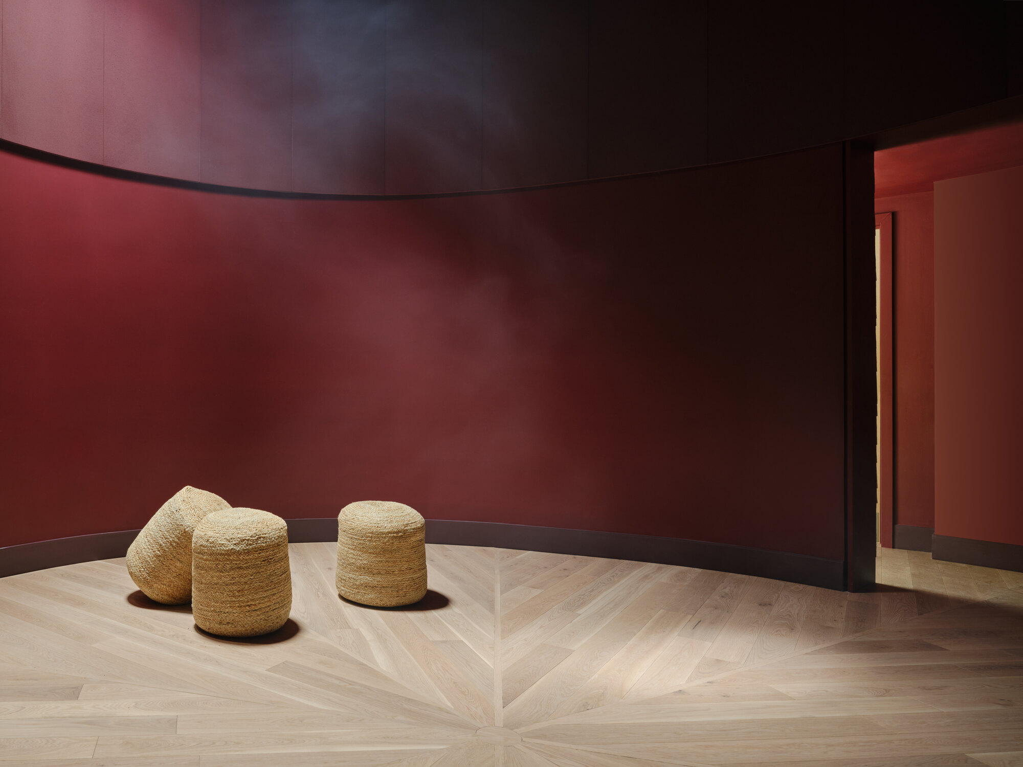 Curved oxblood-red wall enclosing three woven straw pouffes on a pale floor with a radiating herringbone parquet, lit by a soft haze