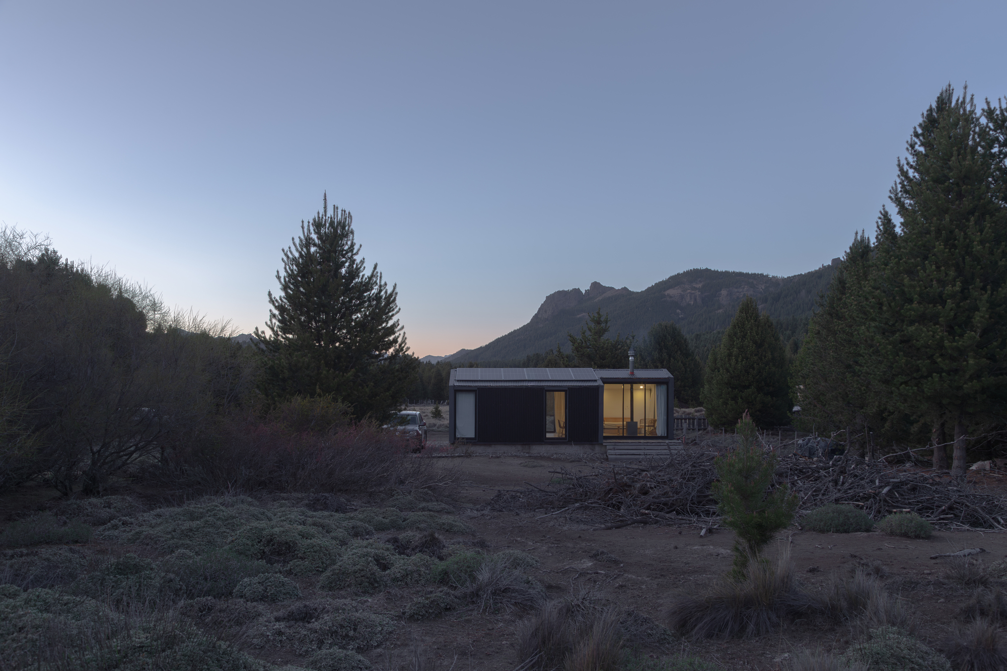 Small dark-clad cabin with a lit glazed end, set among pines and scrub below a rocky ridge at dusk
