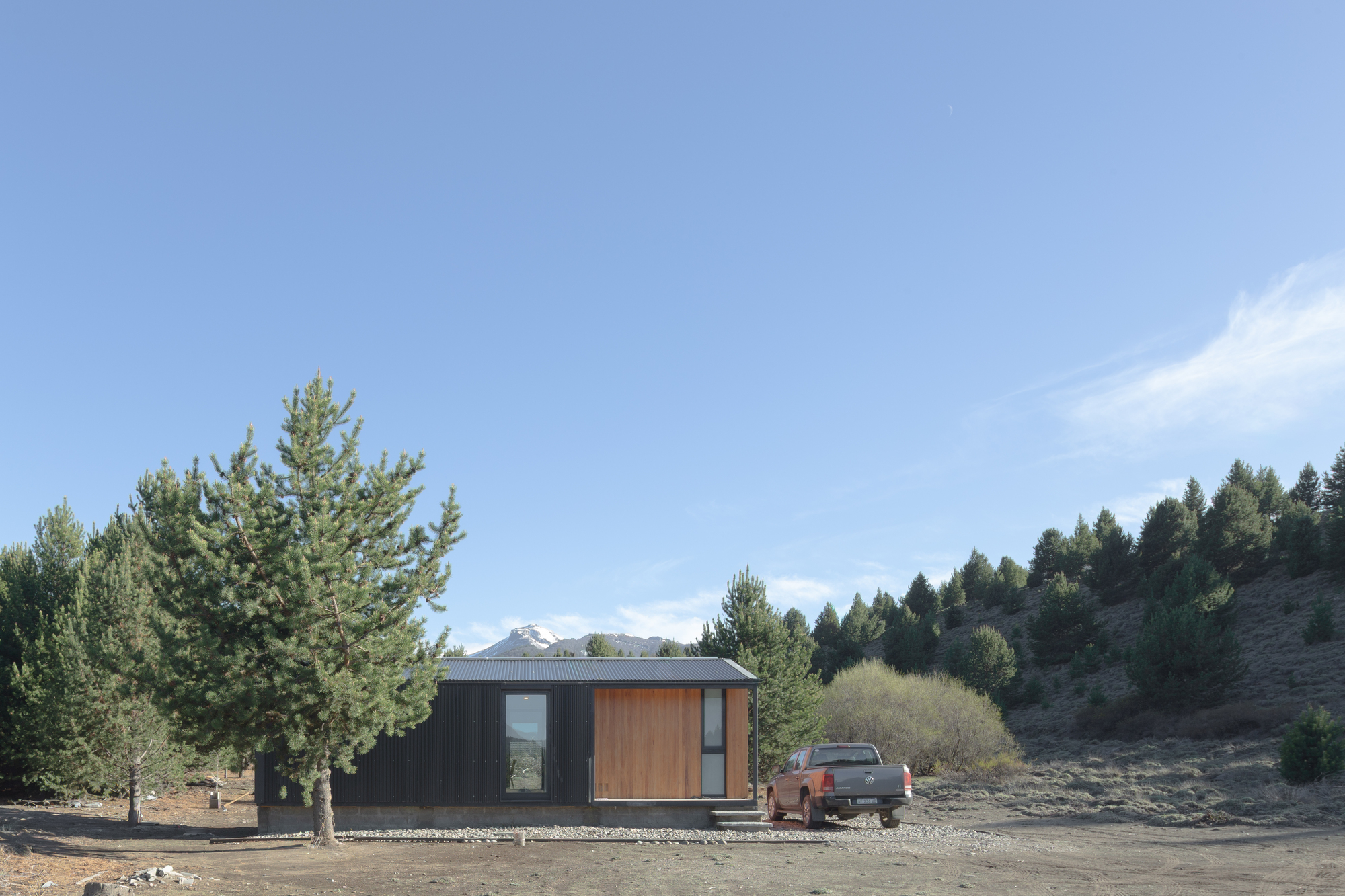 Side elevation of the black corrugated cabin with a timber-clad panel, a pickup parked beside it under a clear blue sky