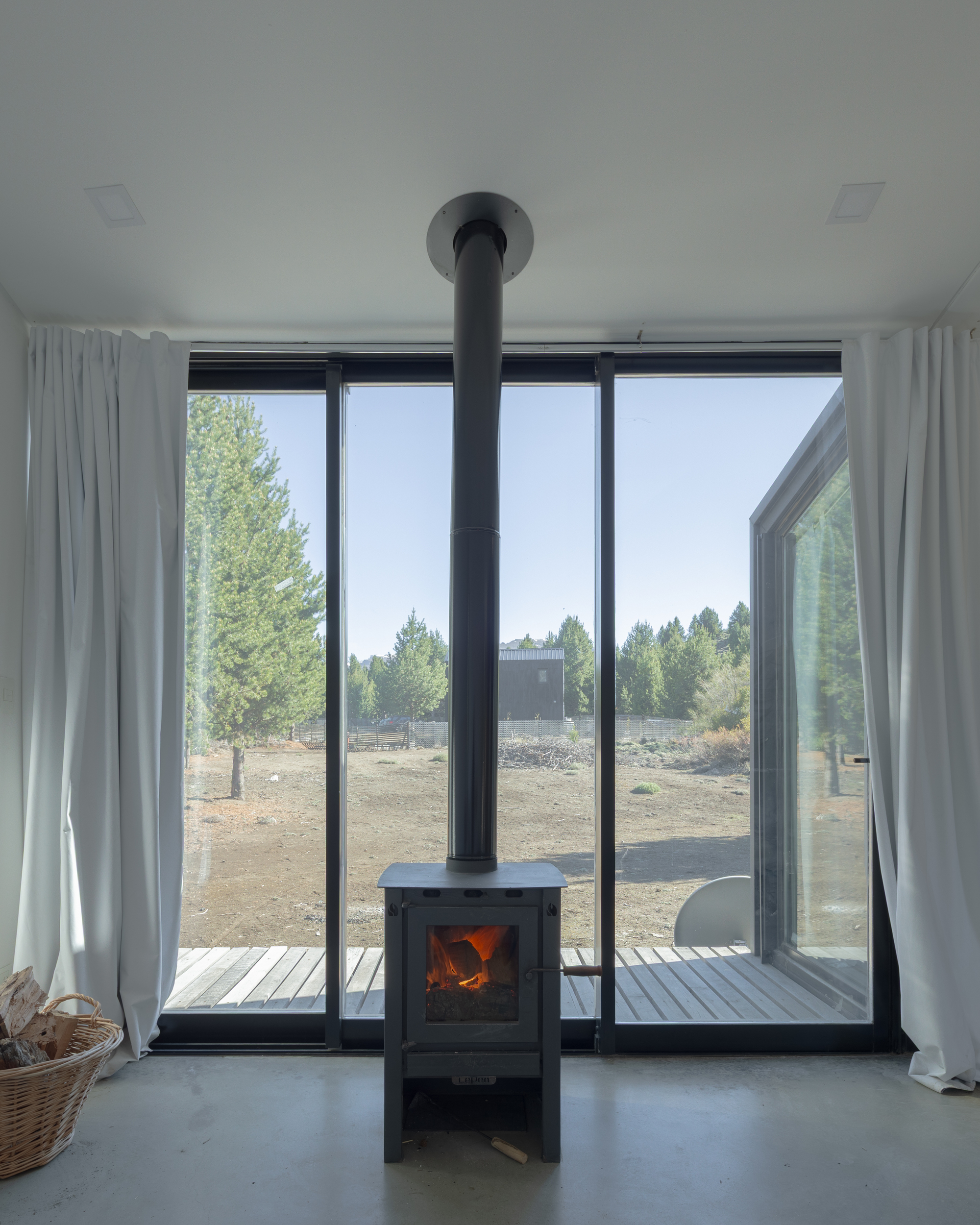 Black wood stove with a vertical flue before a floor-to-ceiling glazed wall, white curtains framing a timber deck and pine trees