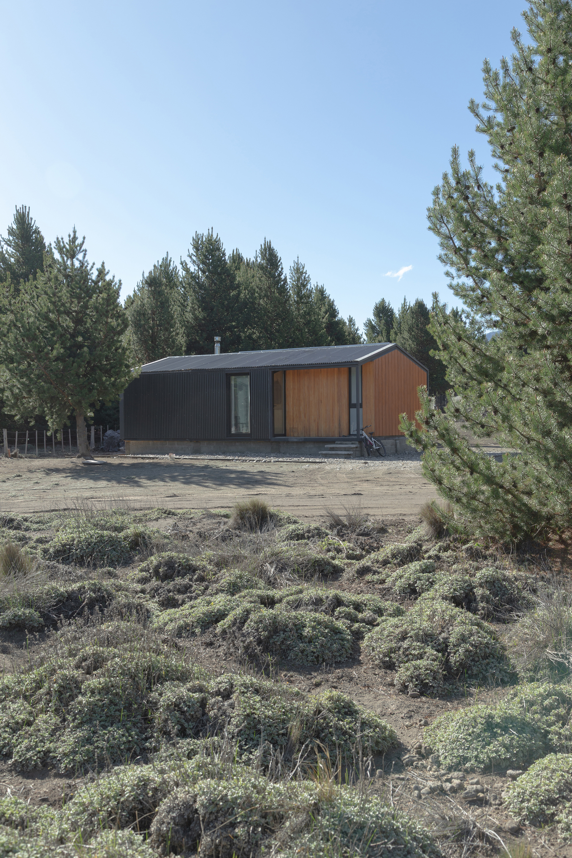 Distant view of the dark cabin with timber end walls behind mounds of low cushion shrubs, a bicycle leaning at its steps
