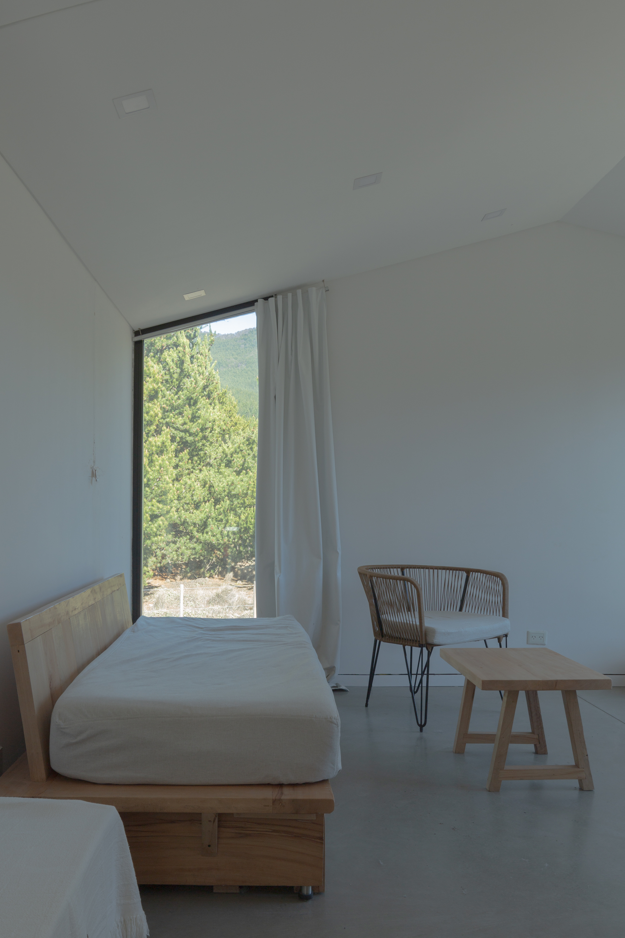 Bedroom with a timber daybed, rope chair and low wooden table beside a tall corner window and white curtain