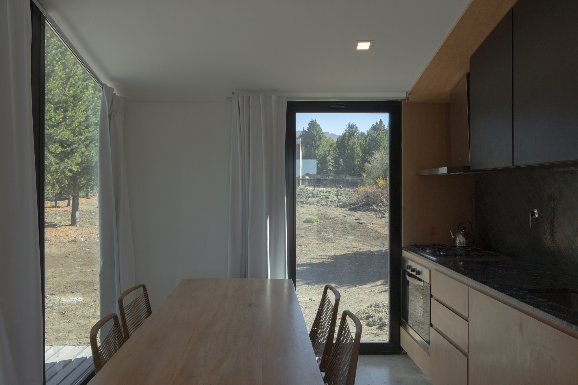 Timber dining table beside a tall black-framed window and kitchen run, with white curtains and pine trees outside in bright sun