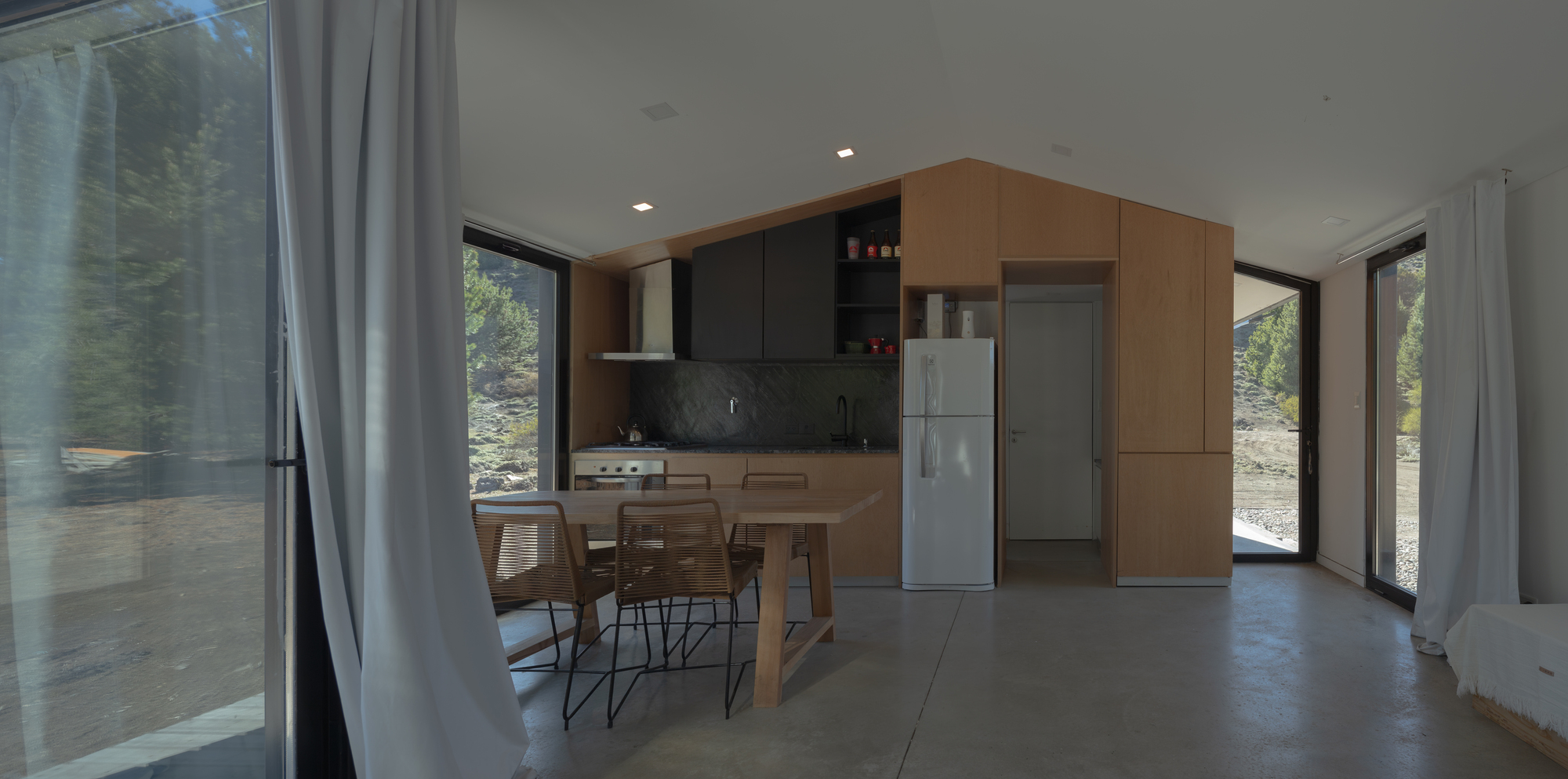 Open living space with a plywood kitchen volume, white fridge and dining table, framed by sheer curtains and sunlit glazing