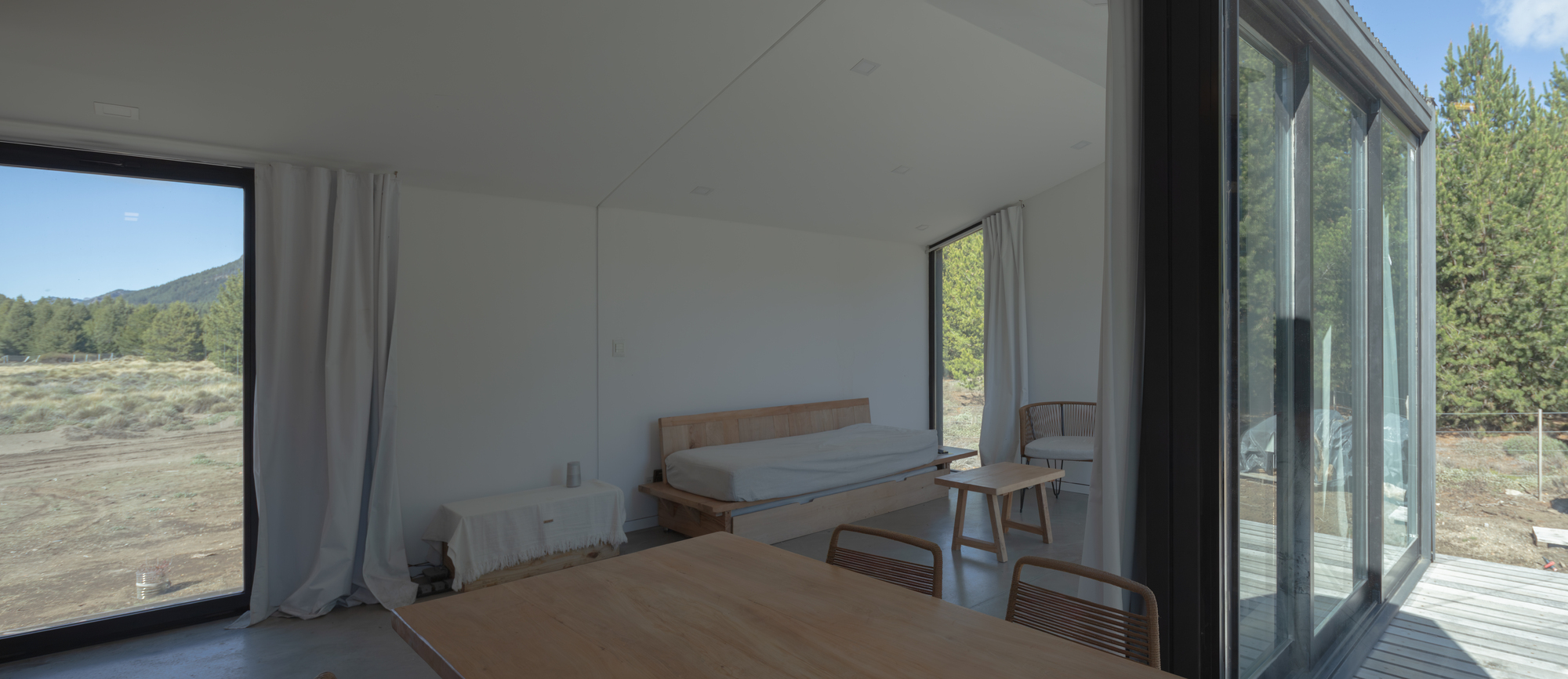 Daybed and small side tables in a white room, with sliding glass doors opening to a timber deck and pines