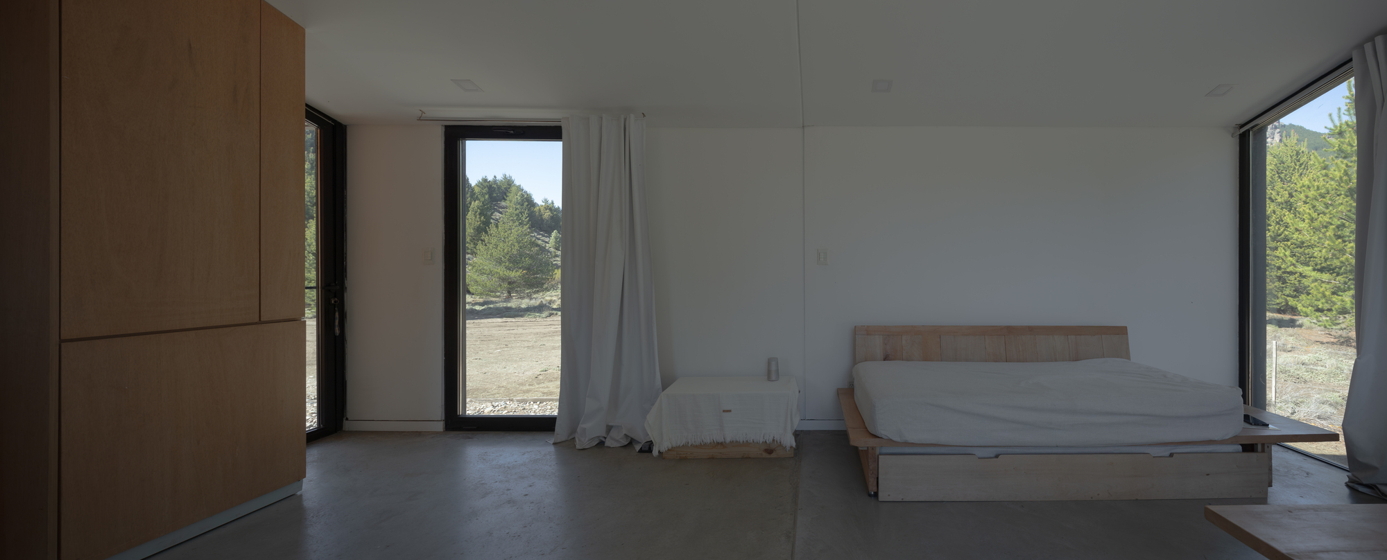 Sleeping area with a low timber daybed and plywood cabinet wall, daylight entering through tall glazed openings onto open grassland