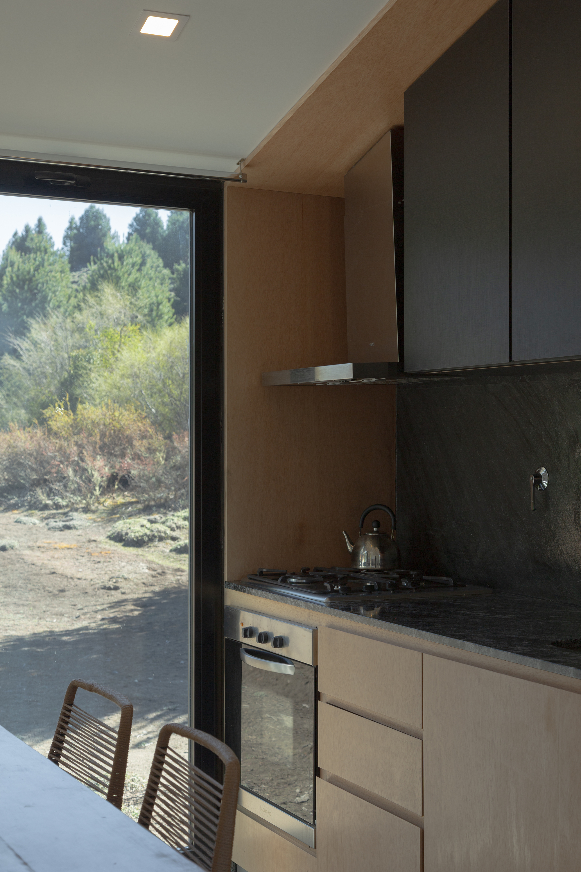 Kitchen corner with dark stone backsplash, gas hob, steel kettle and plywood cabinets beside a window onto autumn shrubs