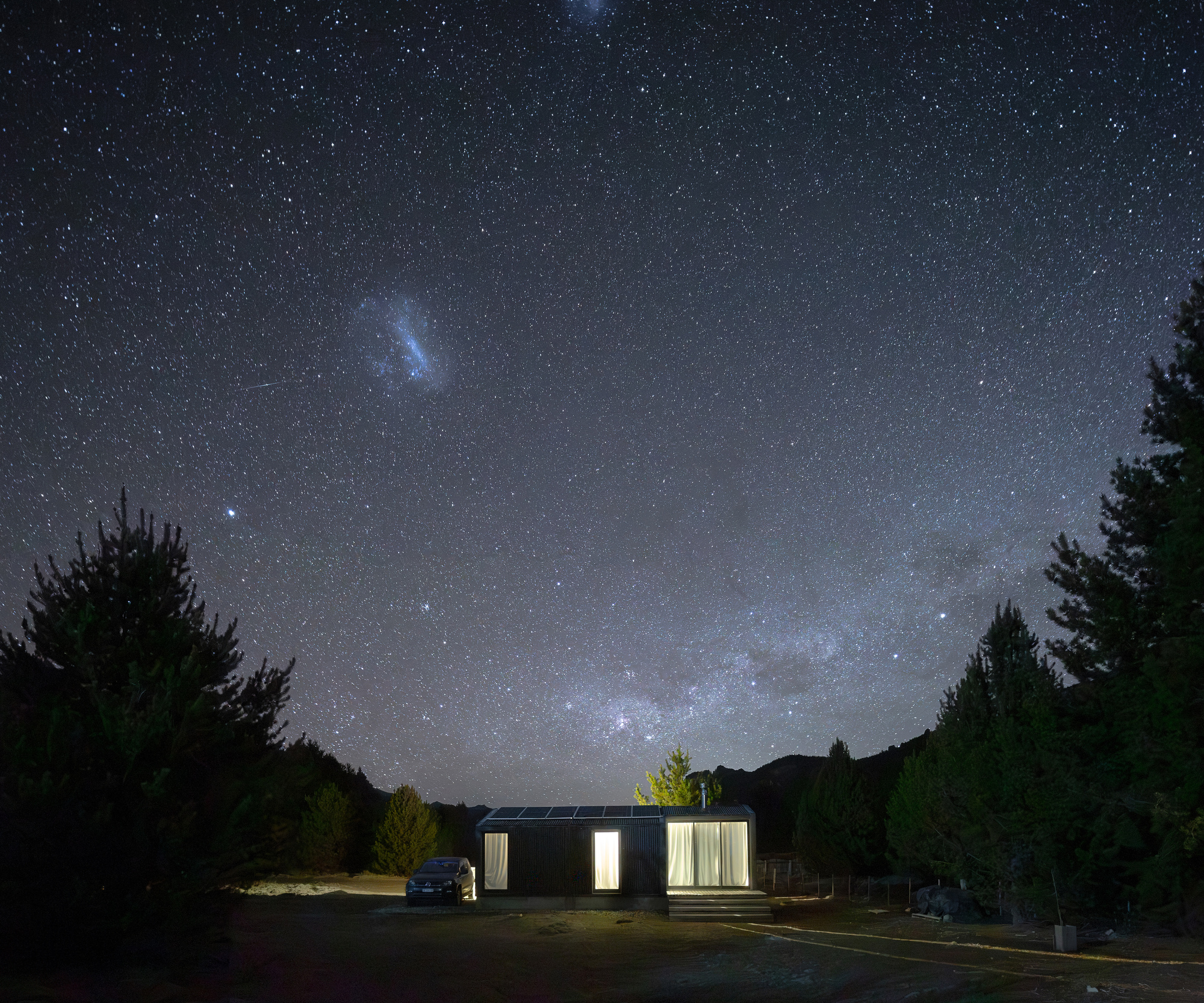 Small dark house with glowing curtained windows and a parked car beneath a star-filled night sky and the Milky Way