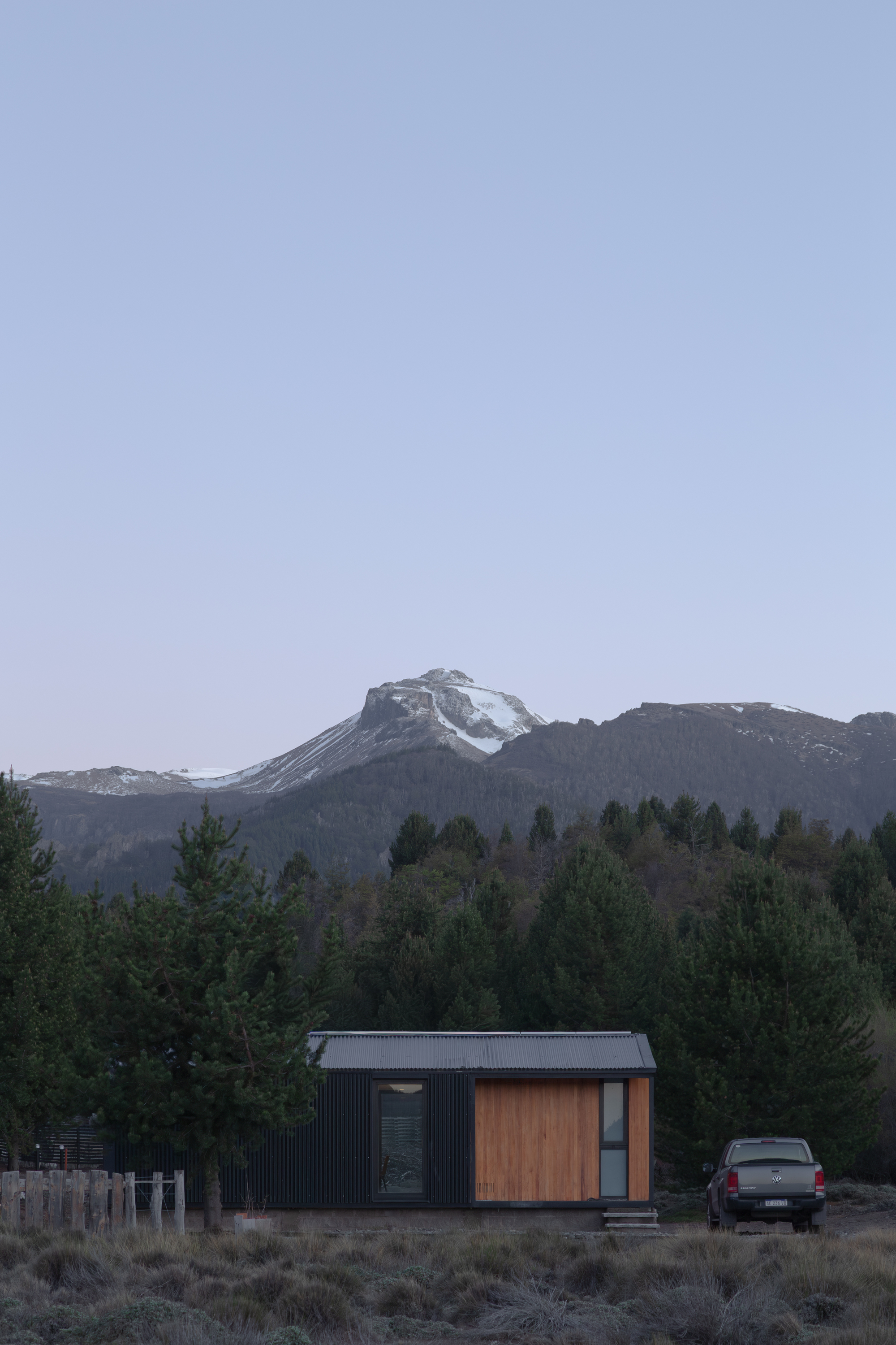 Small cabin with dark cladding and a timber panel below a snow-dusted peak at dusk, a pickup parked beside it
