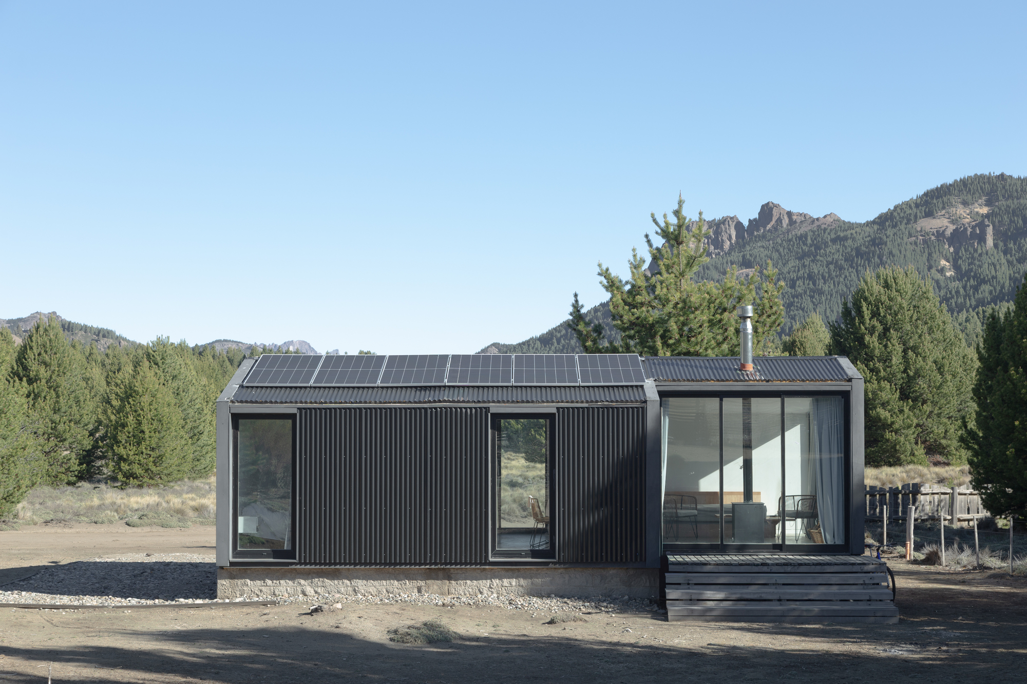 Front elevation of the corrugated black cabin with rooftop solar panels, a glazed living end and a timber deck step in sunlight