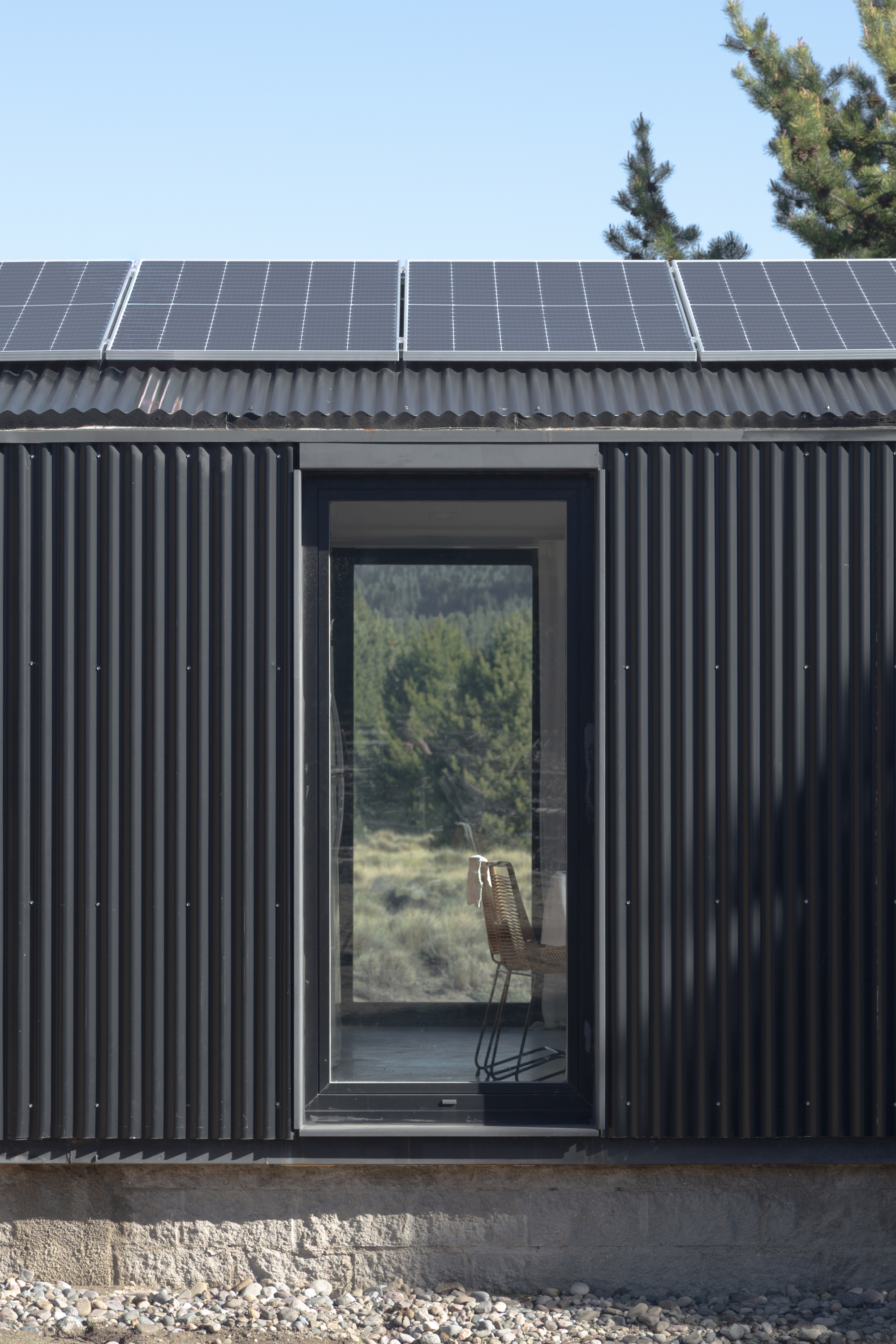 Close-up of the tall window set in corrugated black cladding beneath solar panels, with a woven chair visible inside