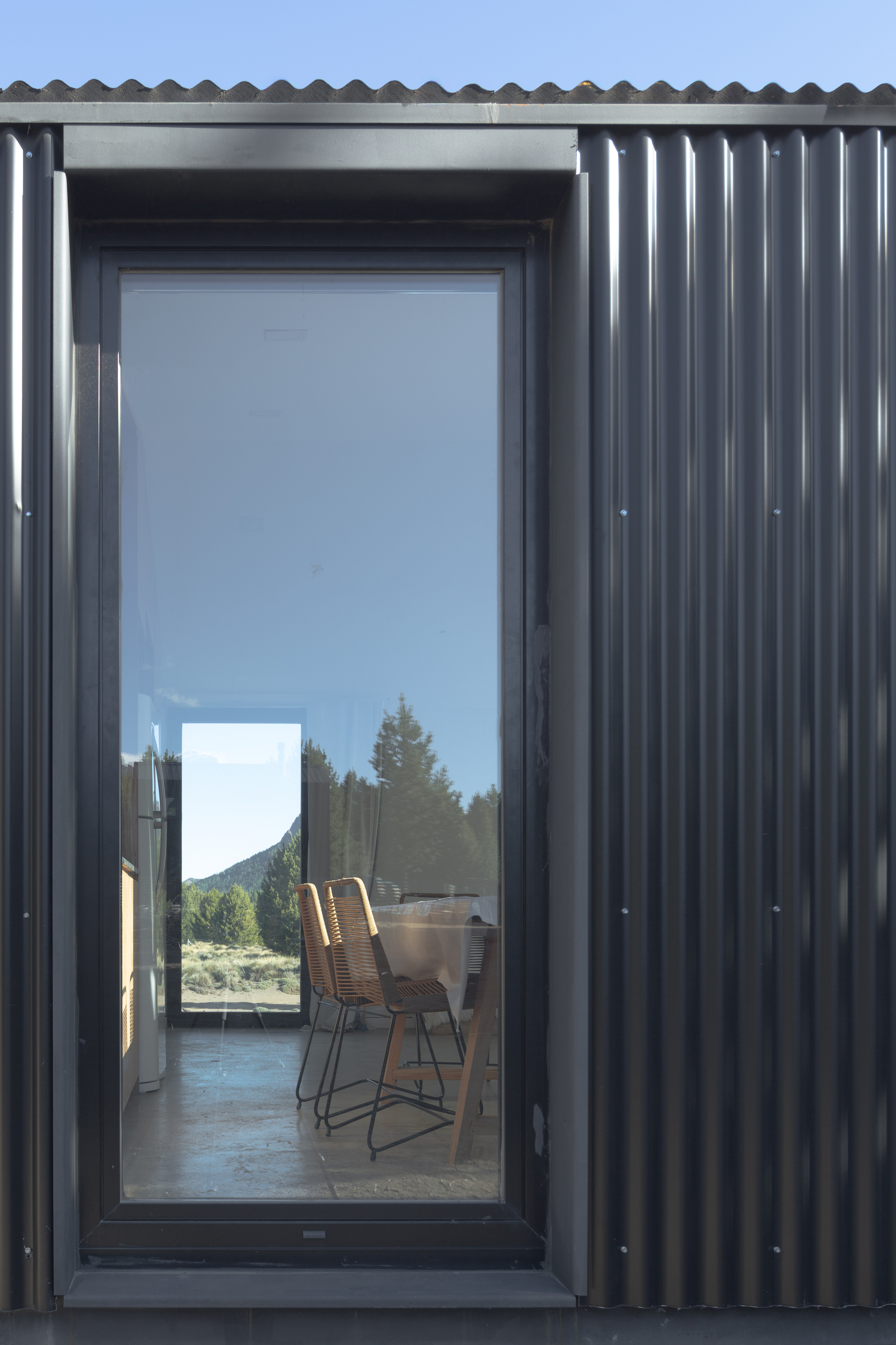 Detail of a tall window in corrugated metal cladding reflecting pines and a mountain, with woven chairs inside