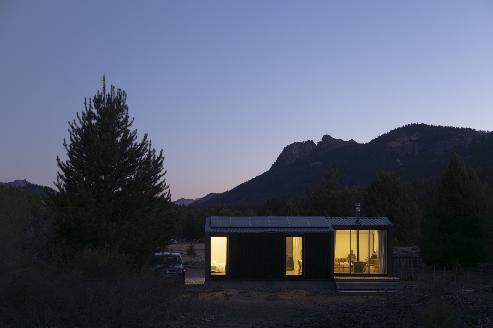 Small cabin with glowing windows and a solar-panel roof at dusk, a pine tree and rocky ridge behind