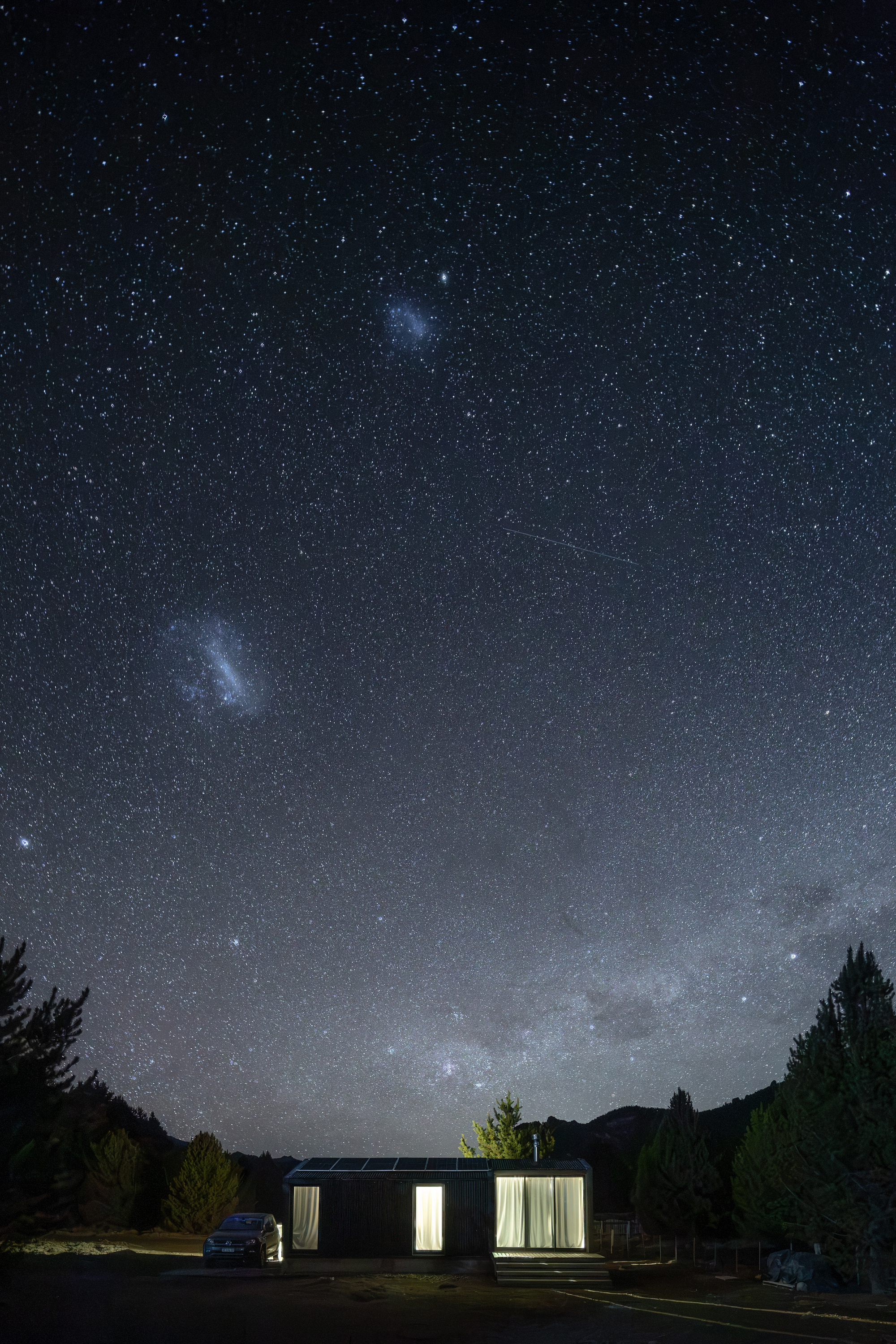 Lit cabin beneath a dense starry night sky, with two faint galaxies above the pines and a parked car