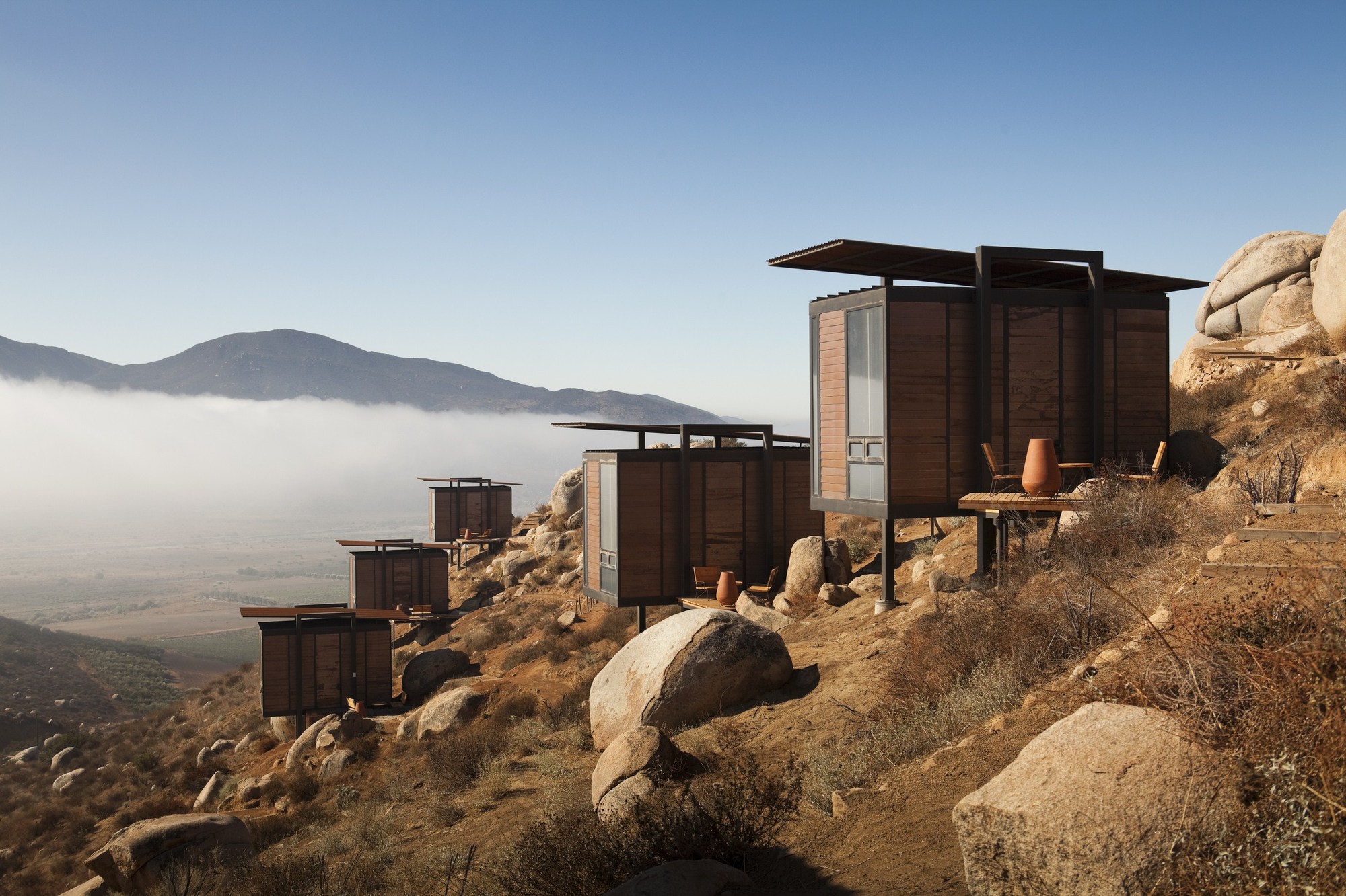 Row of timber-clad cabins on stilts along a rocky ridge, with low morning fog filling the valley behind