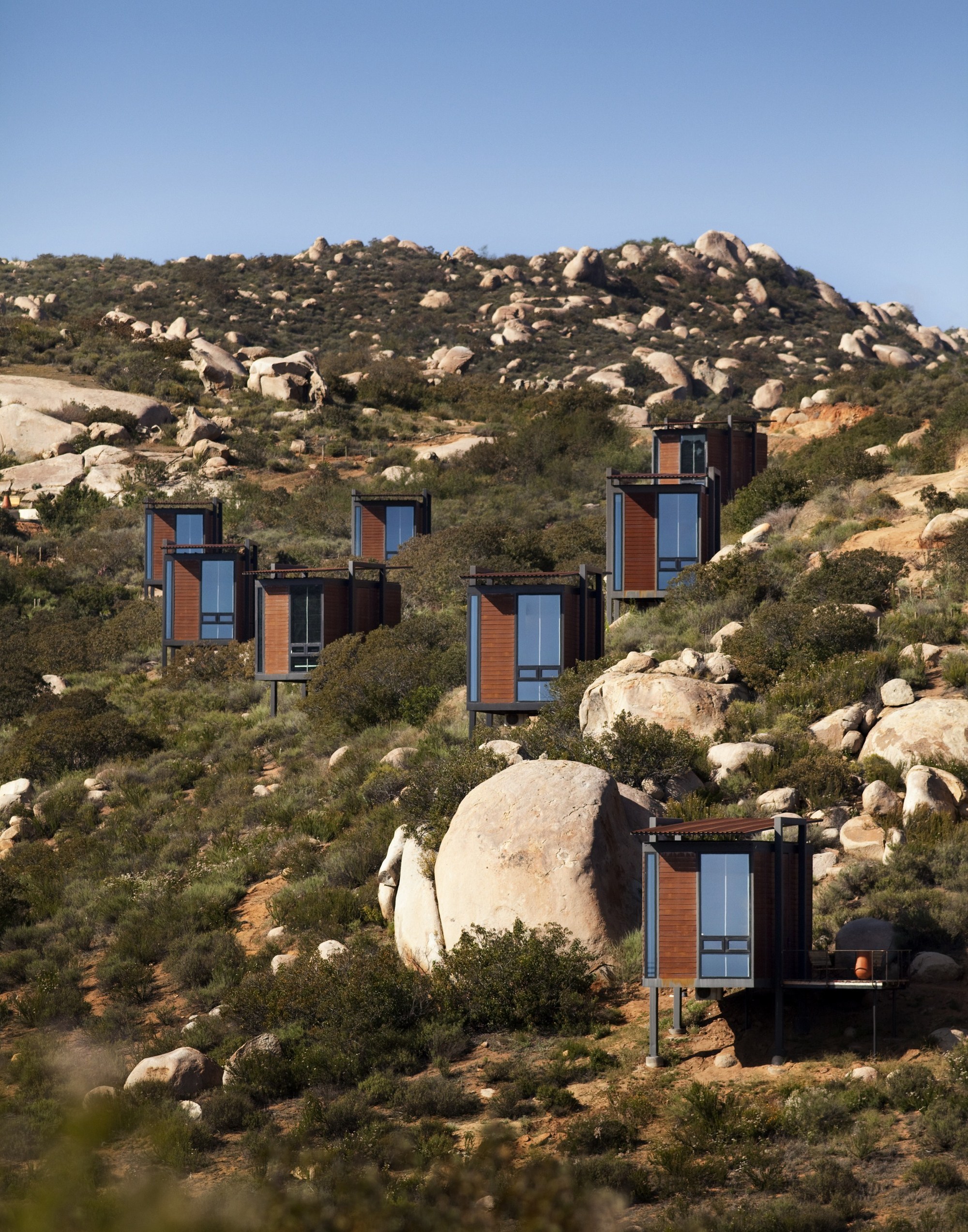 Staggered timber cabins with glazed ends climbing a boulder-strewn hillside under a clear blue sky