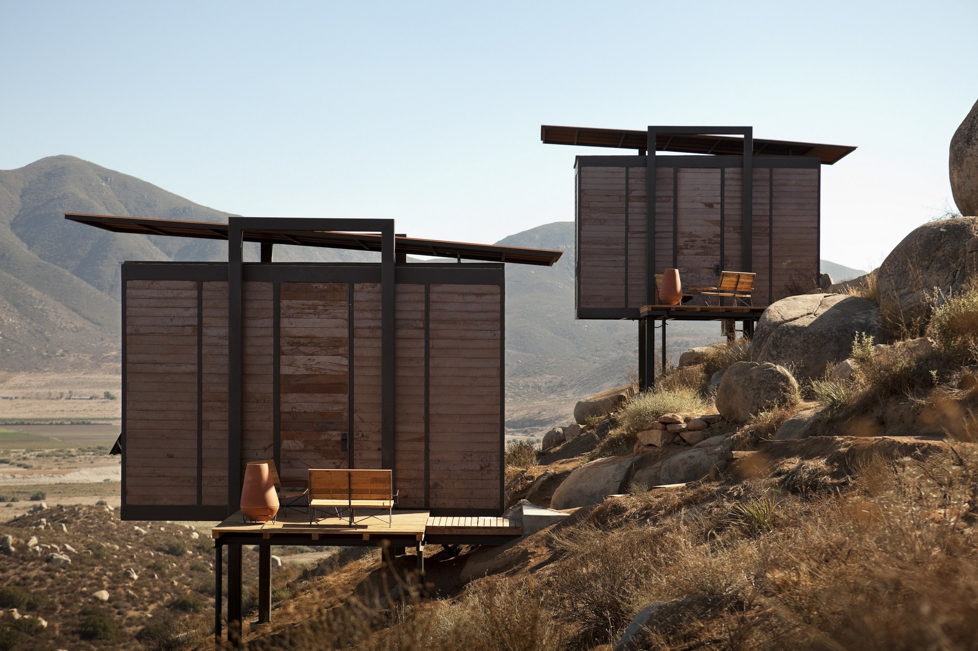 Two raised cabins with slatted timber walls, steel frames and thin cantilevered roofs, seen in warm afternoon sun