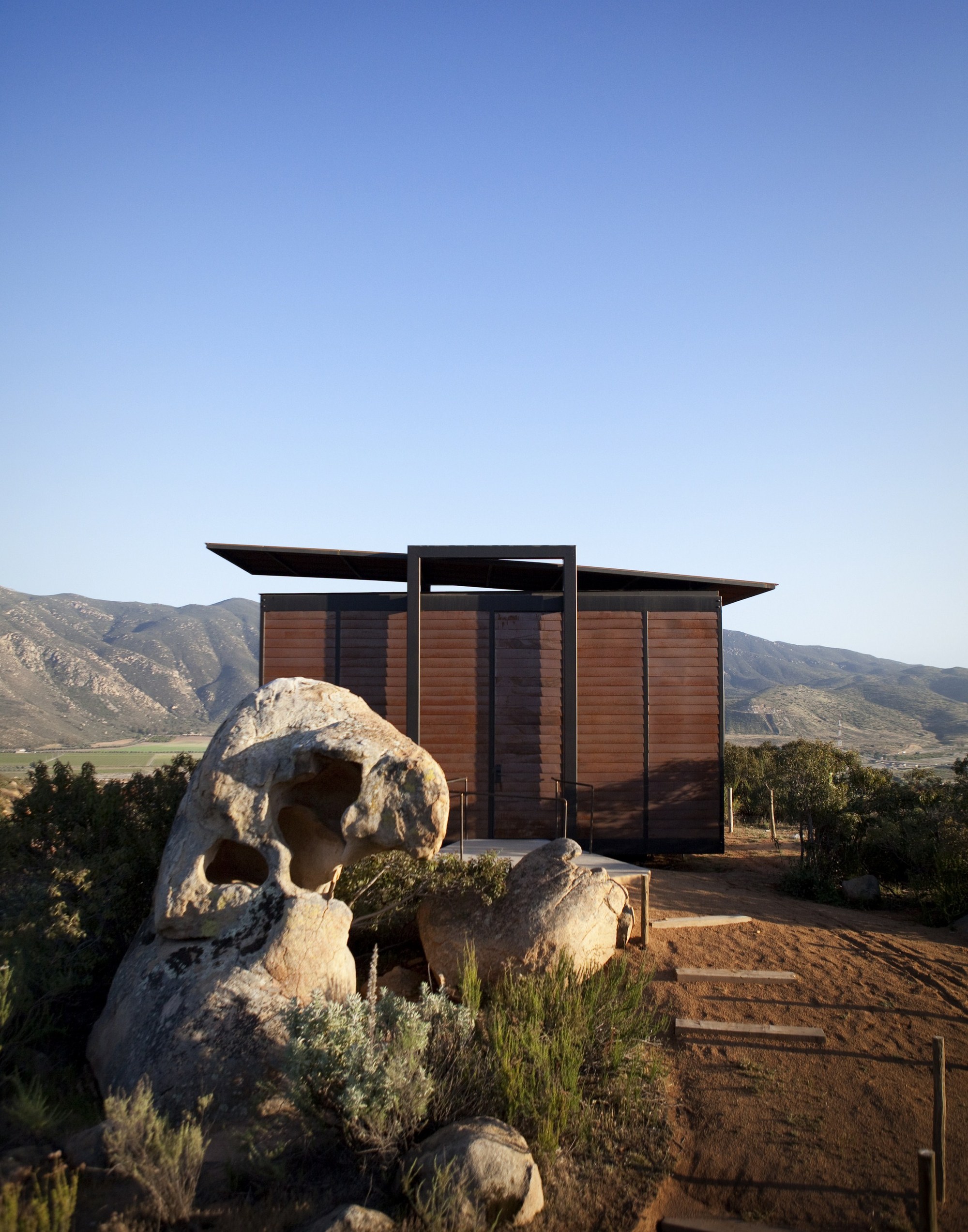 Front view of a timber-clad cabin with a floating roof plane behind a hollowed granite boulder at dusk