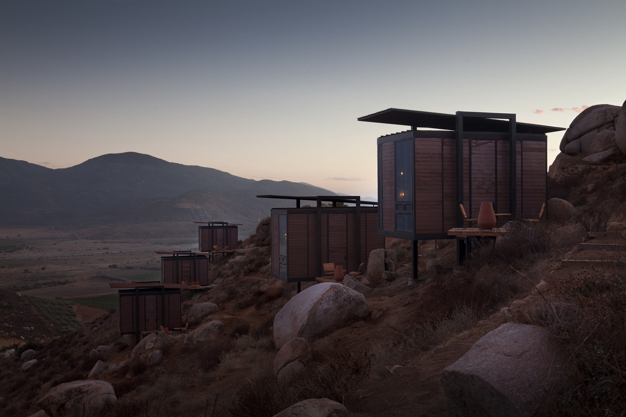 Cabins perched on the slope at twilight, one window glowing warmly against dark mountains and a pink sky