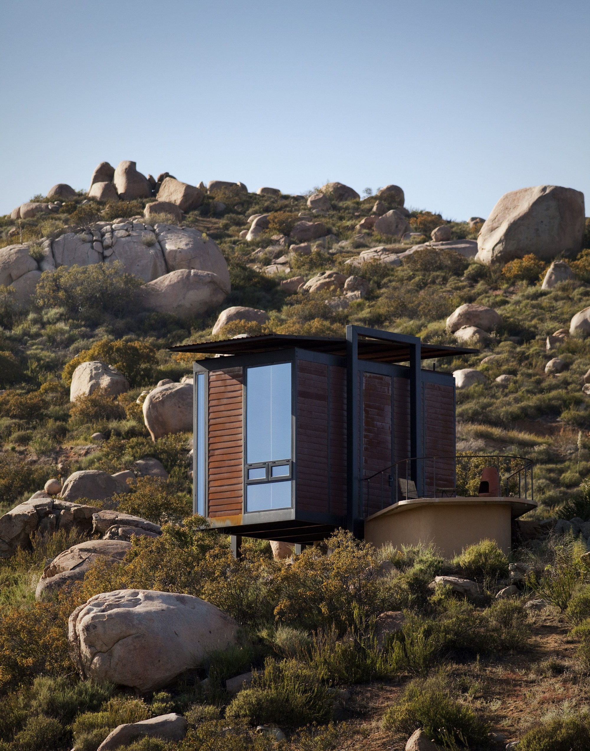 Single cabin with a tall glazed corner and slatted timber walls, set among boulders and low scrub in afternoon light