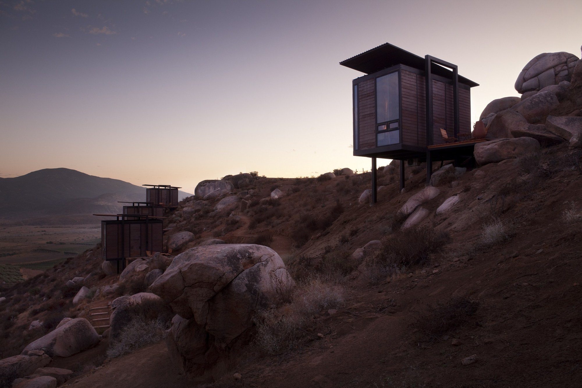 Timber-clad cabin raised on steel stilts above boulders, with a row of cabins stepping down the slope at dusk