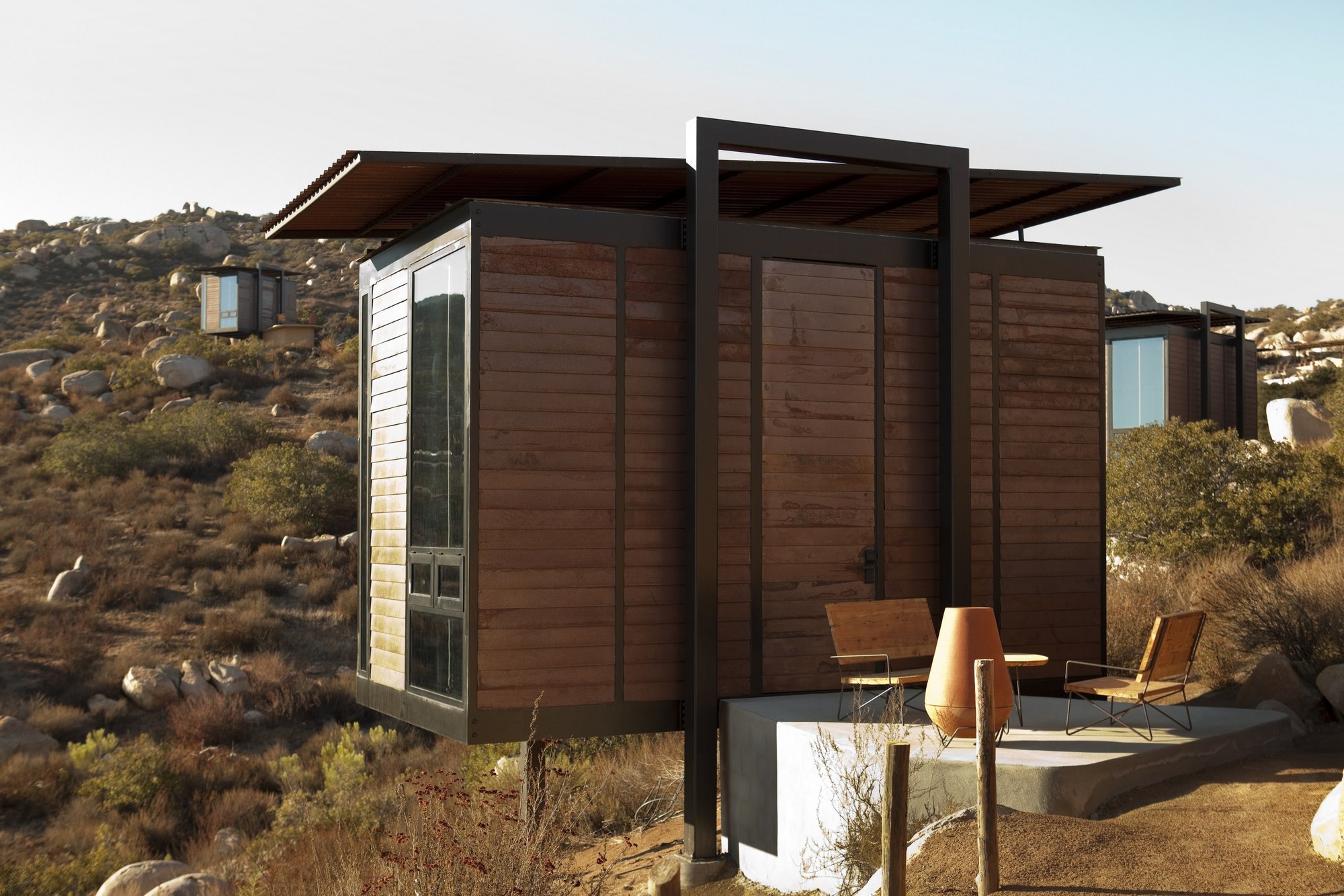 Wood-slatted cabin with a cantilevered steel-framed roof and a concrete patio holding chairs and a clay fireplace in low sun