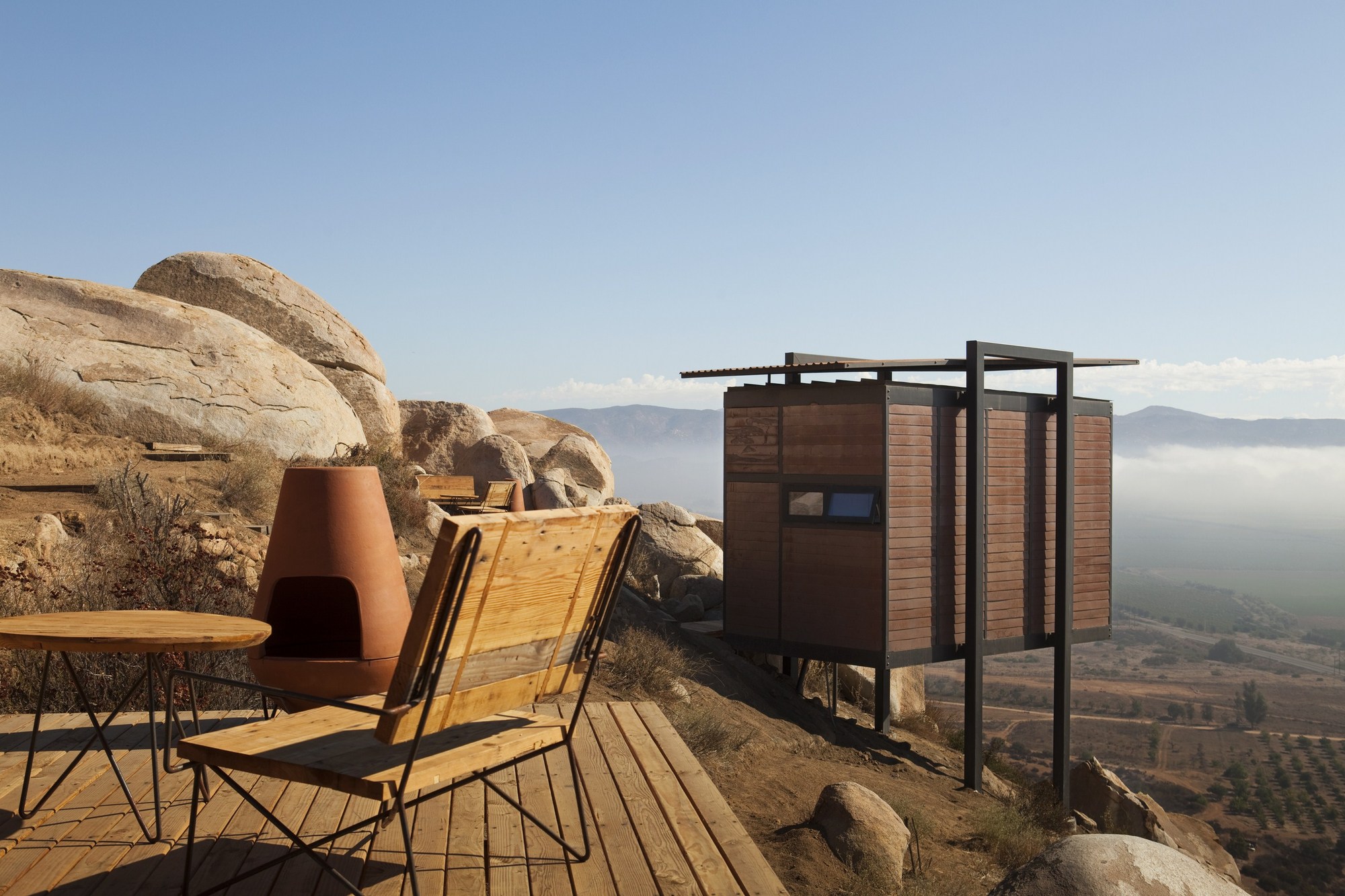 Wooden deck with a lounge chair, round table and terracotta fireplace facing a stilted cabin above a hazy valley