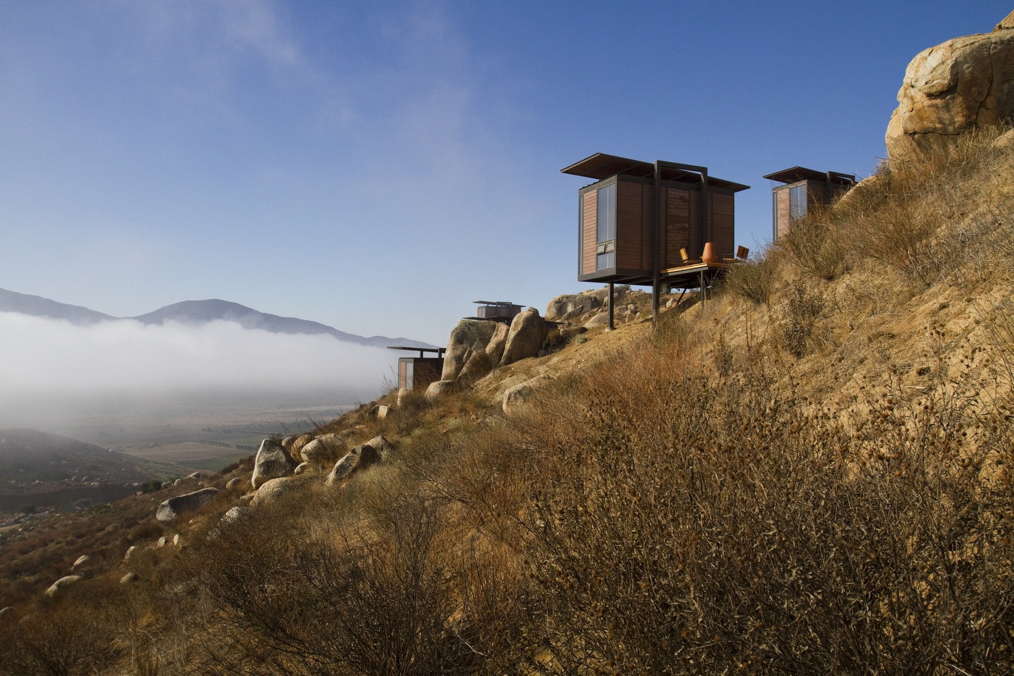 Two stilted timber cabins perched on a dry hillside above a bank of low fog in the valley
