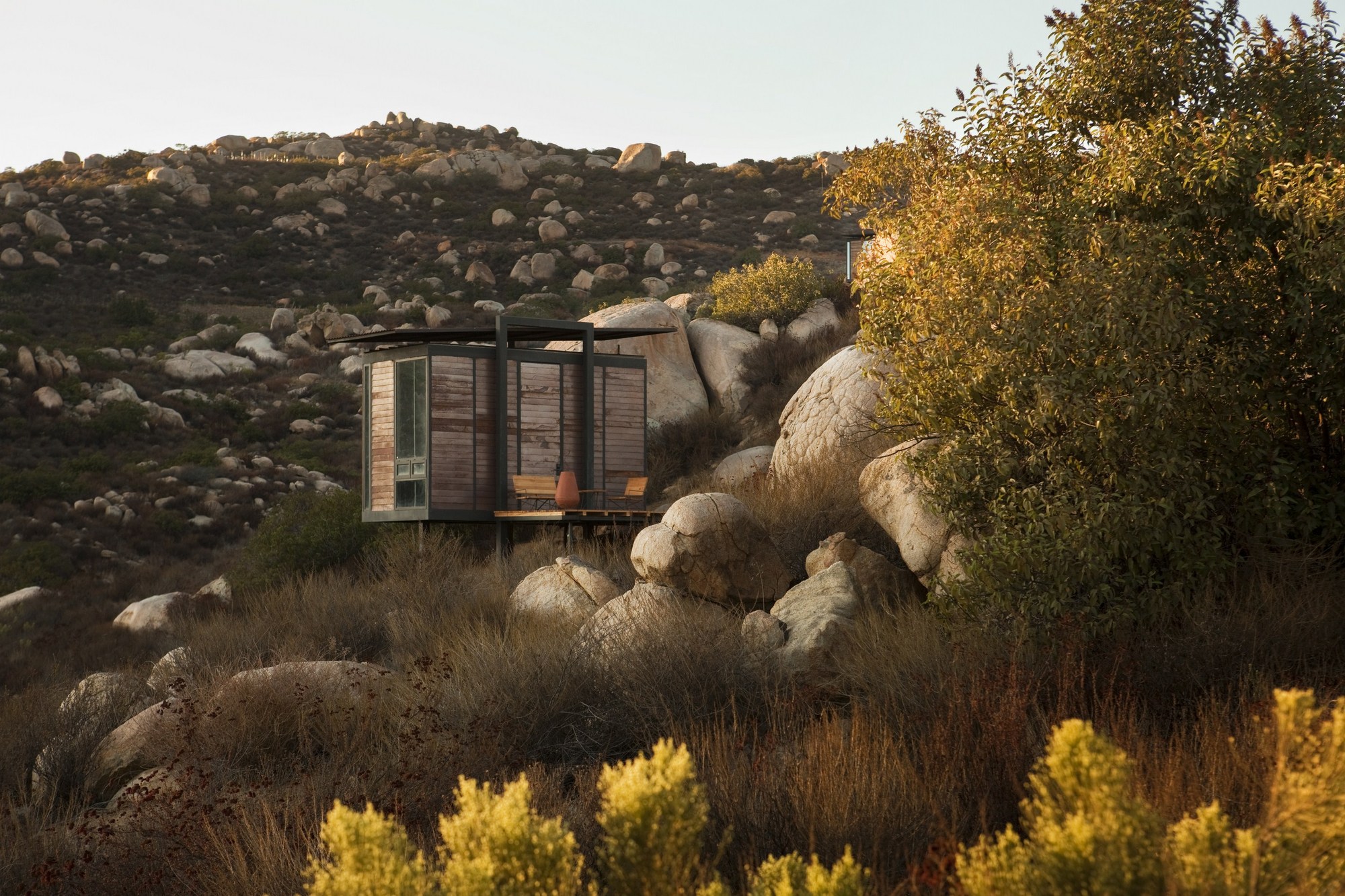 Glazed cabin with slatted timber panels nestled among granite boulders and shrubs in warm late afternoon light