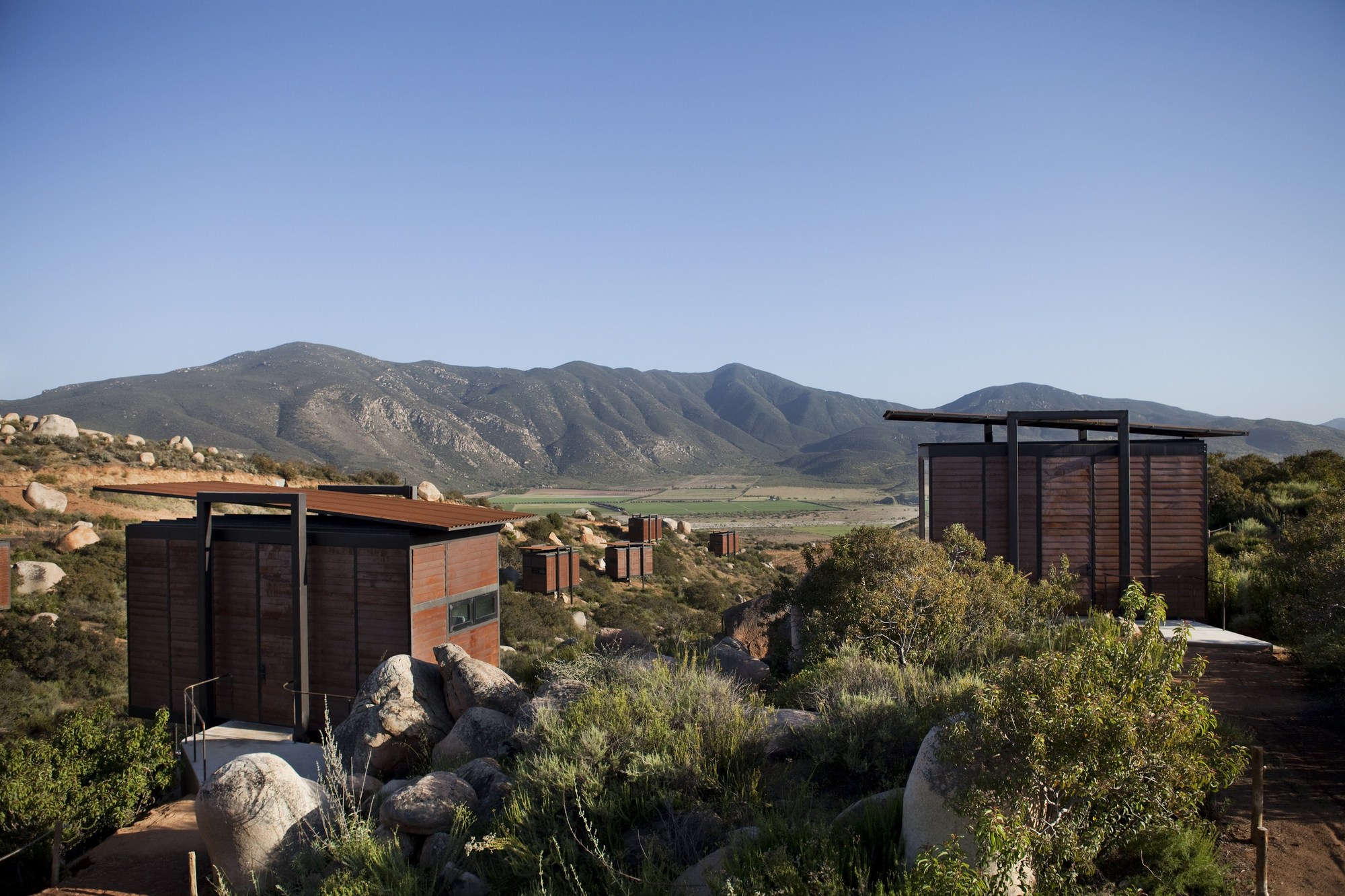Slatted cabins with flat steel roofs scattered across a scrubby hillside, looking toward green fields and distant mountains under clear sky