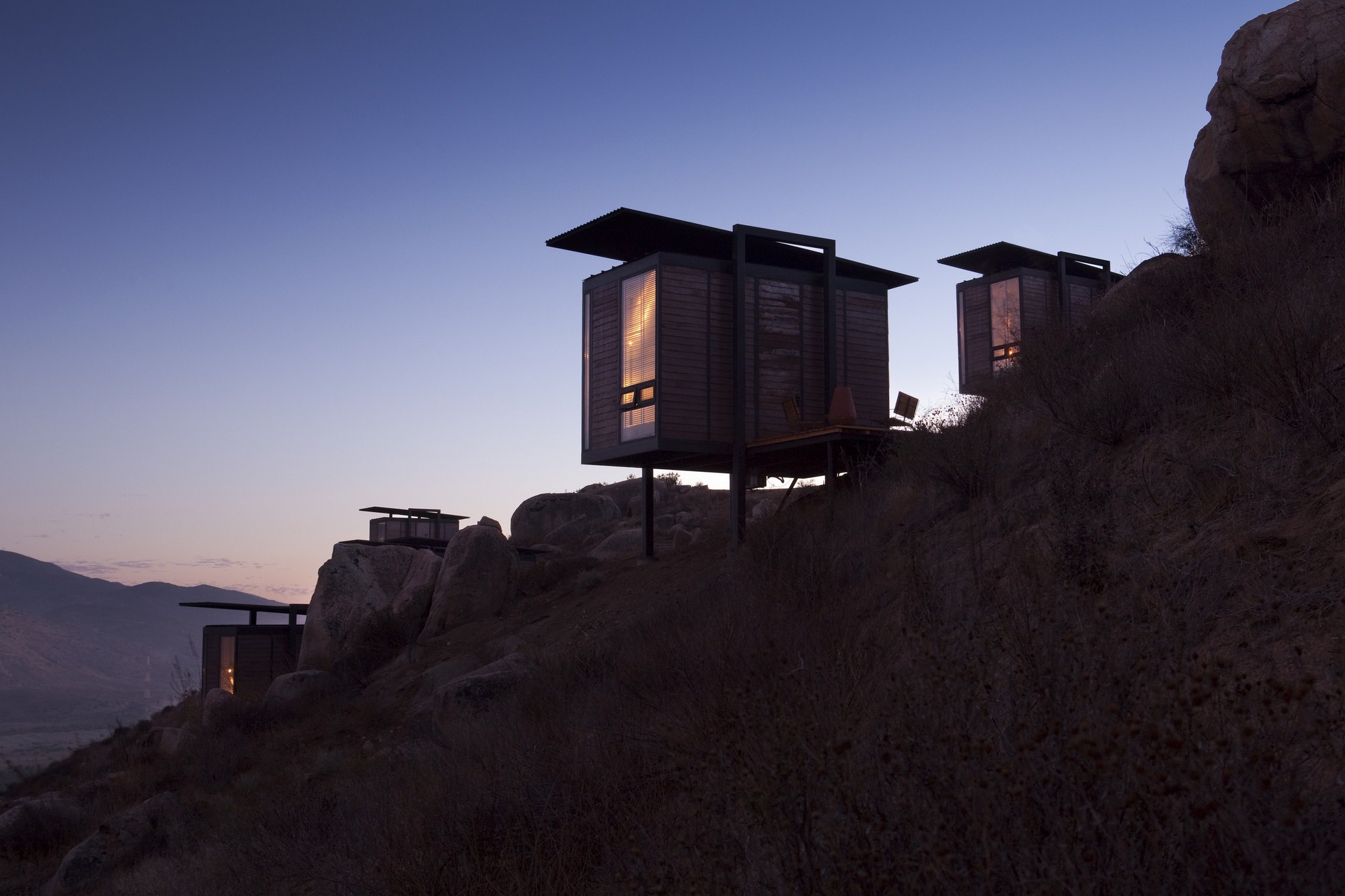 Silhouetted timber-clad cabins on stilts perched among boulders on a hillside, windows glowing at dusk