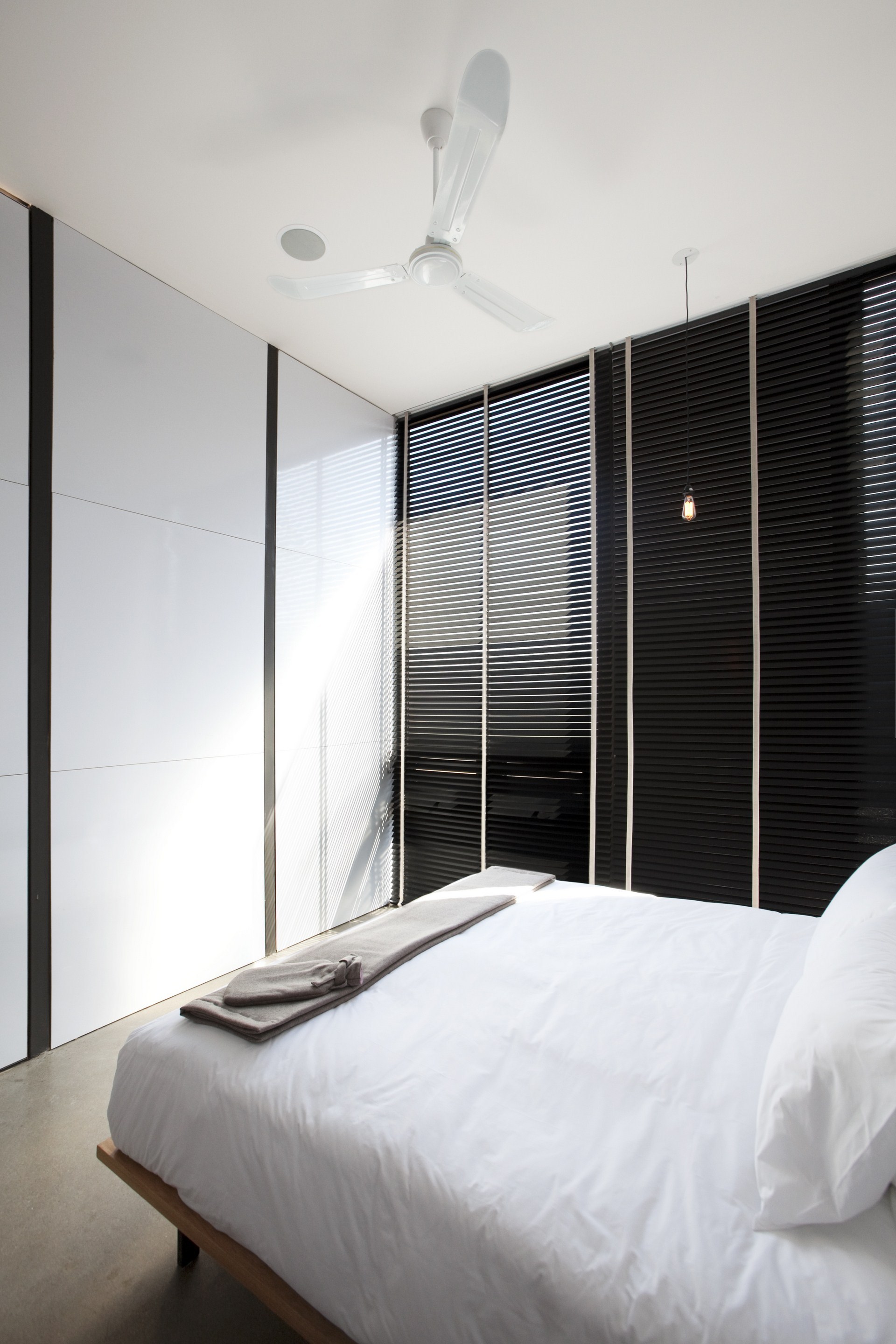 Bedroom corner with white wall panels beside black slatted blinds, a ceiling fan above a low platform bed in sunlight