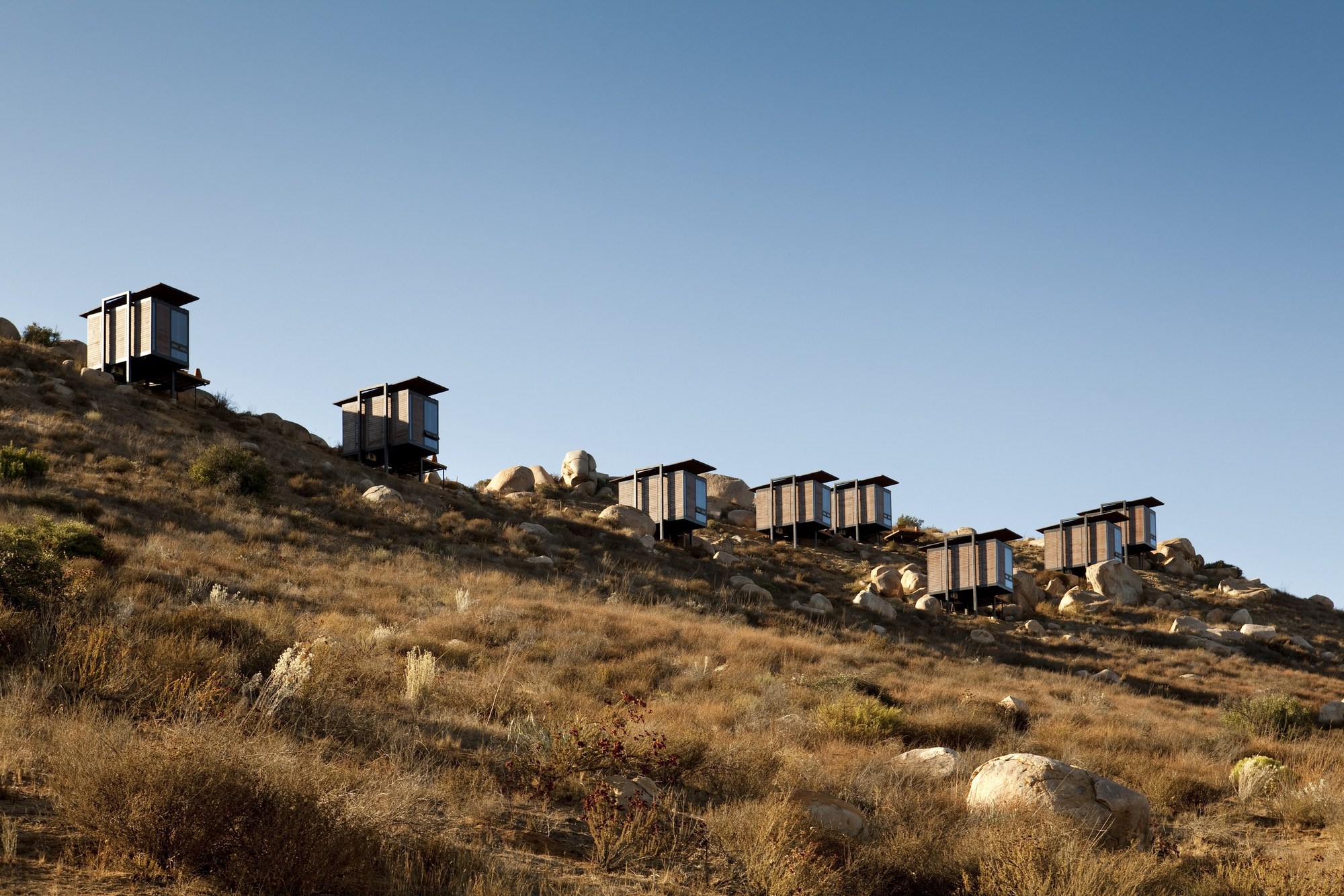 Row of elevated cabins with dark flat roofs stepping along a dry grass hillside under a clear blue sky