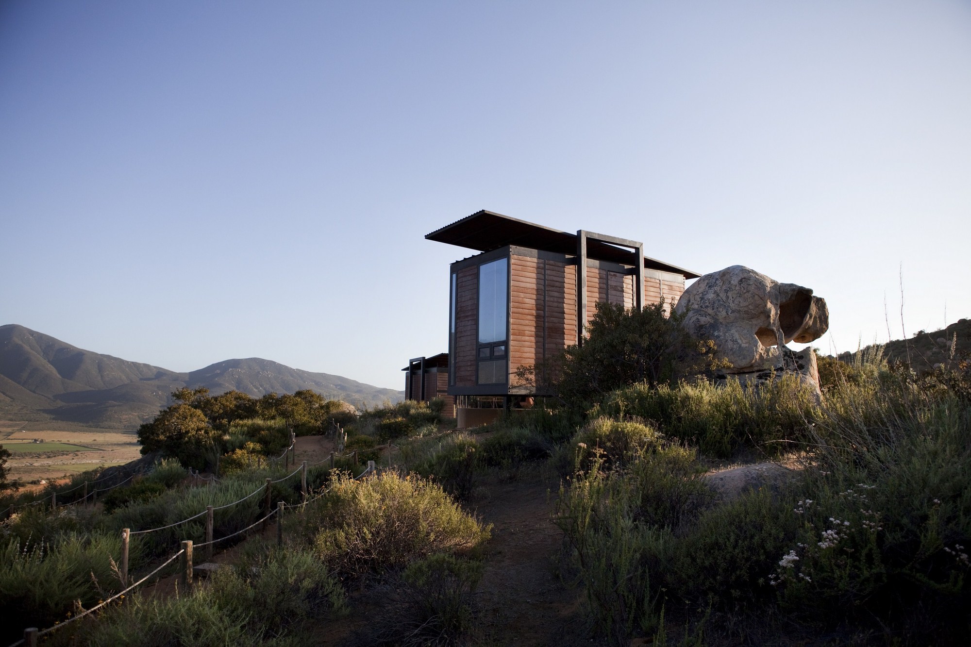 Wood-clad cabin with cantilevered steel roof beside a large boulder, a rope-fenced path winding through shrubs in evening light