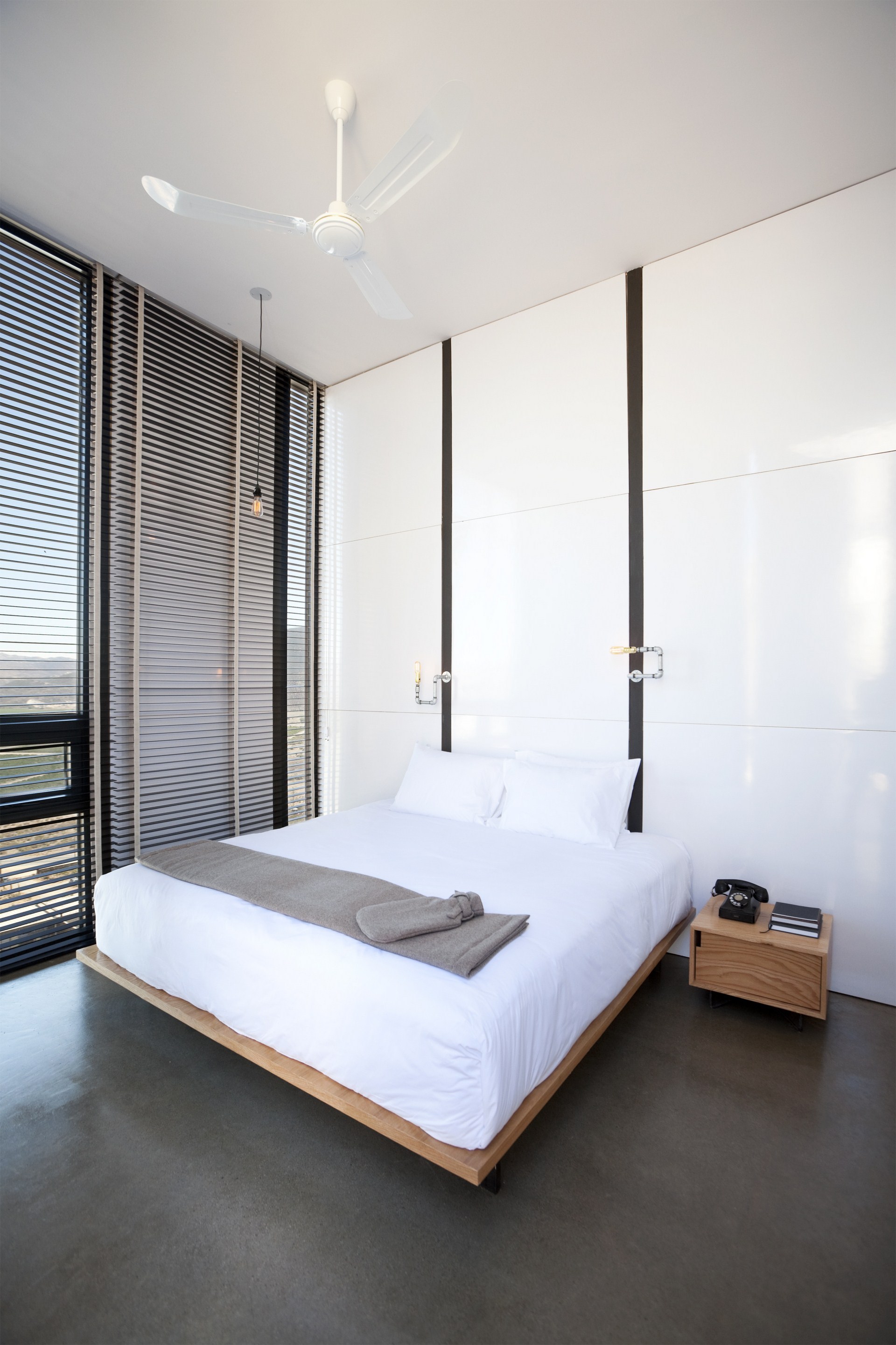 Bedroom with glossy white wall panels, a wood platform bed and a rotary phone on a bedside table, blinds along the window wall