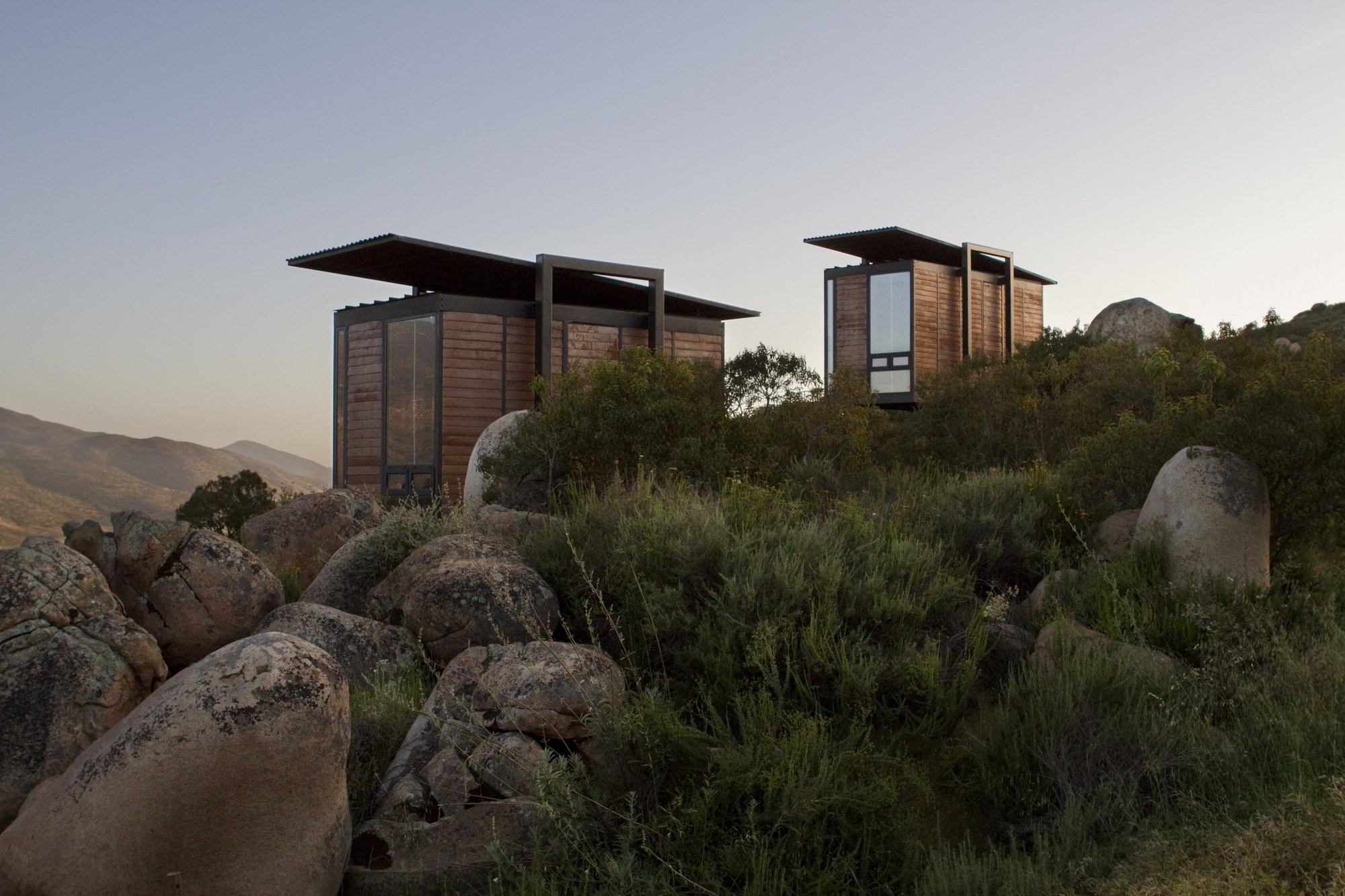 Two timber-clad cabins with thin cantilevered roofs rising above green scrub and rounded granite boulders at dusk